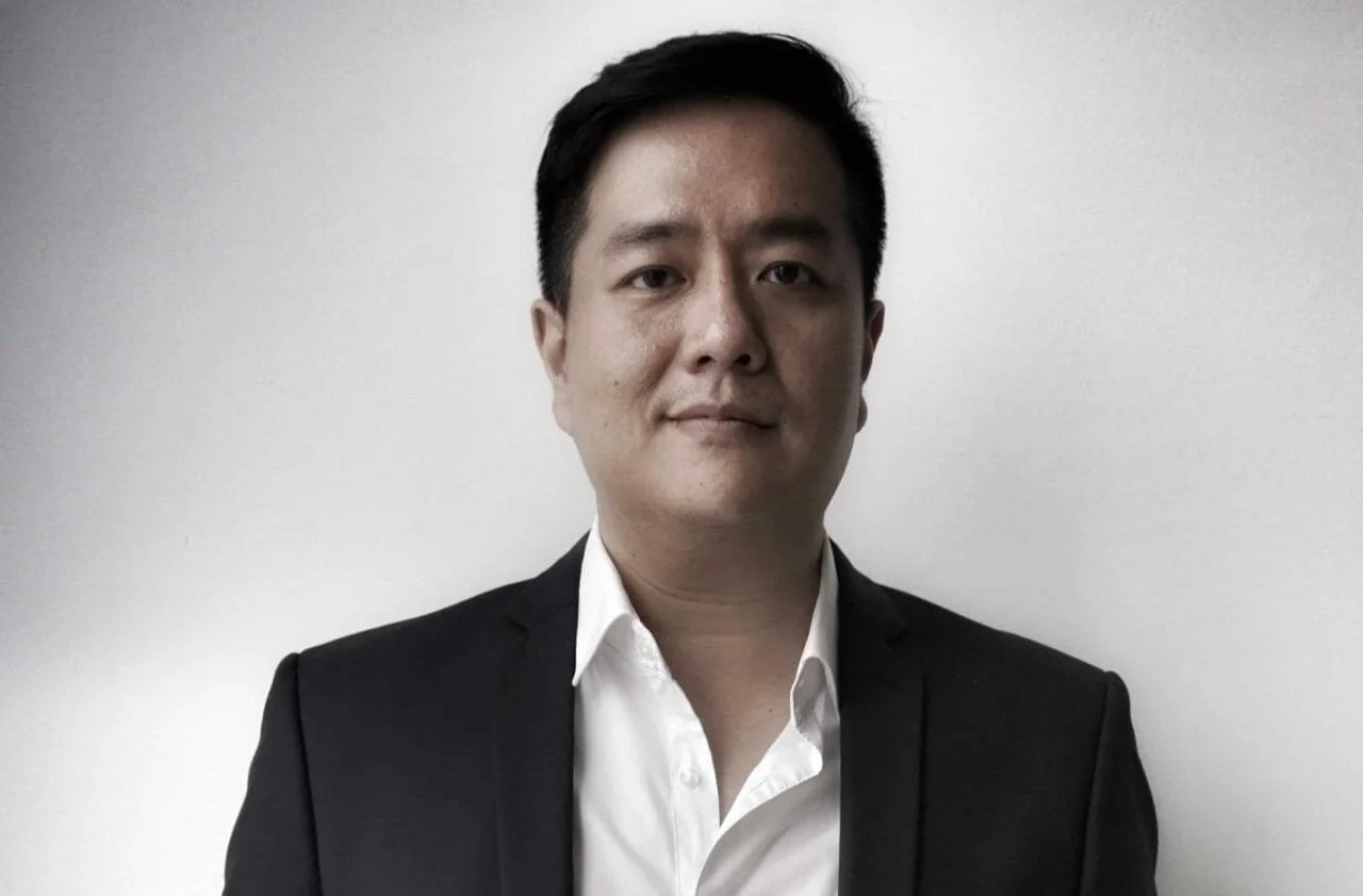 A man in a black suit and white shirt stands against a plain white wall.
