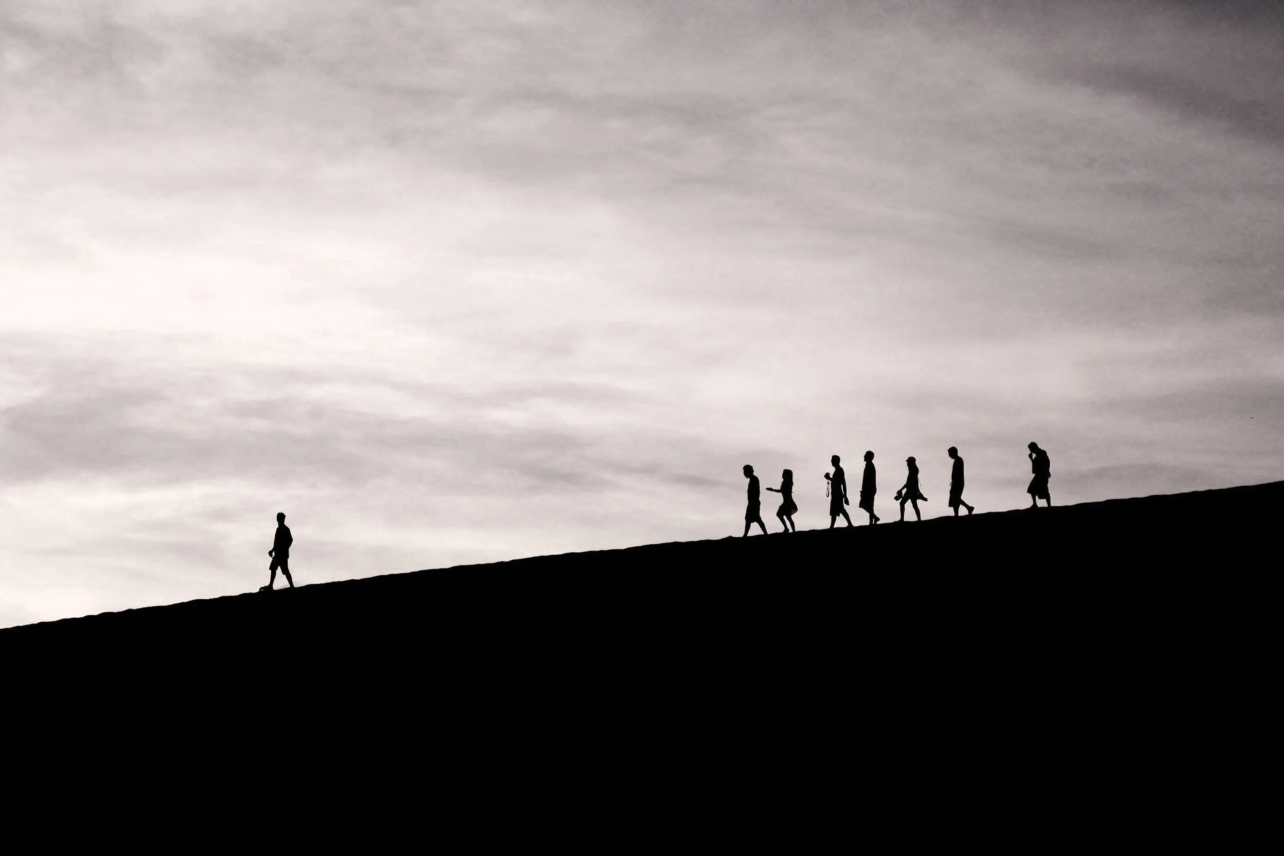 Silhouettes of people walking uphill on a hill at sunset or sunrise with a cloudy sky in the background.