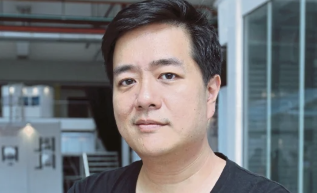 A man with short dark hair and light skin, wearing a black shirt, standing in an indoor space with large windows and modern architecture.