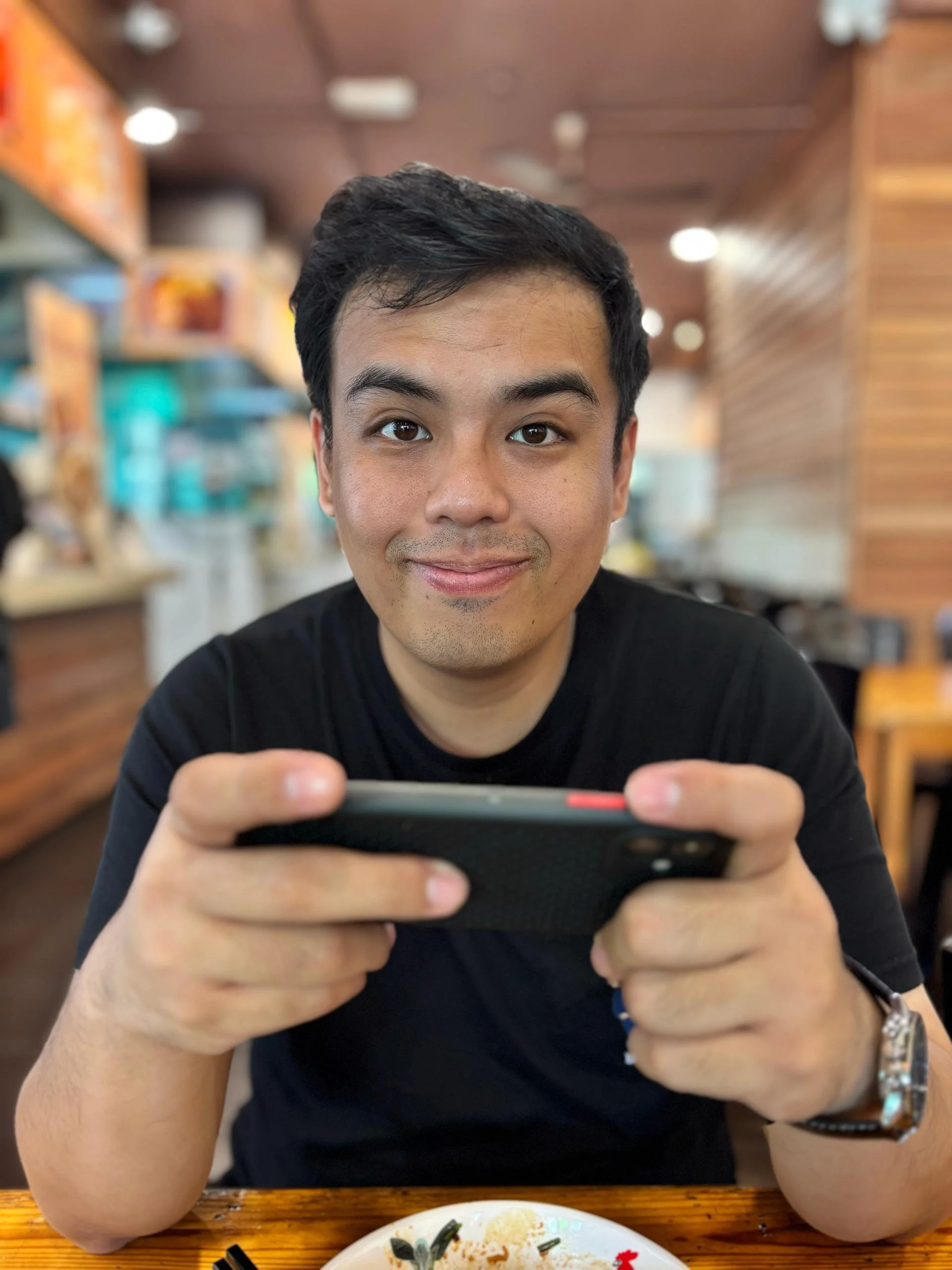A man with short black hair smiling and holding a smartphone while sitting at a restaurant table with a plate of food in front of him. The background features a blurred restaurant interior with wooden accents.