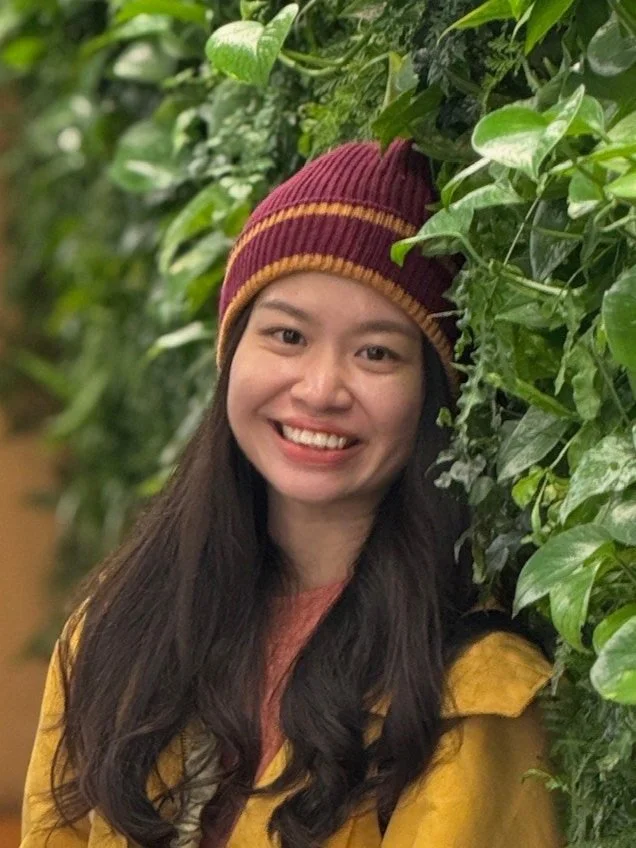 A young woman with long dark hair and a wide smile, wearing a maroon and yellow striped beanie and a mustard yellow jacket, standing next to a lush green leafy wall.