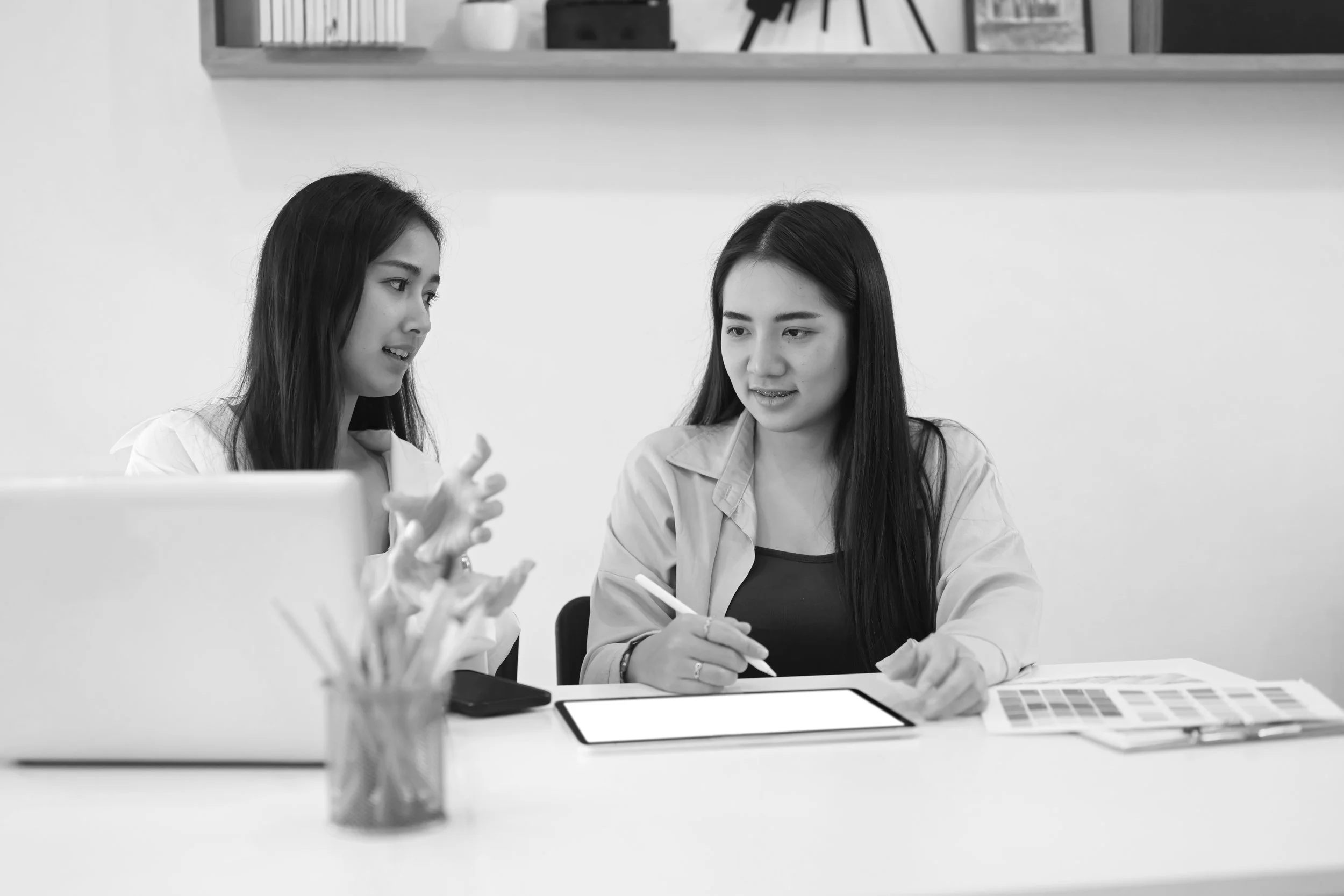 Two women sitting at a table having a discussion, with a laptop, a tablet, and color swatches in front of them.