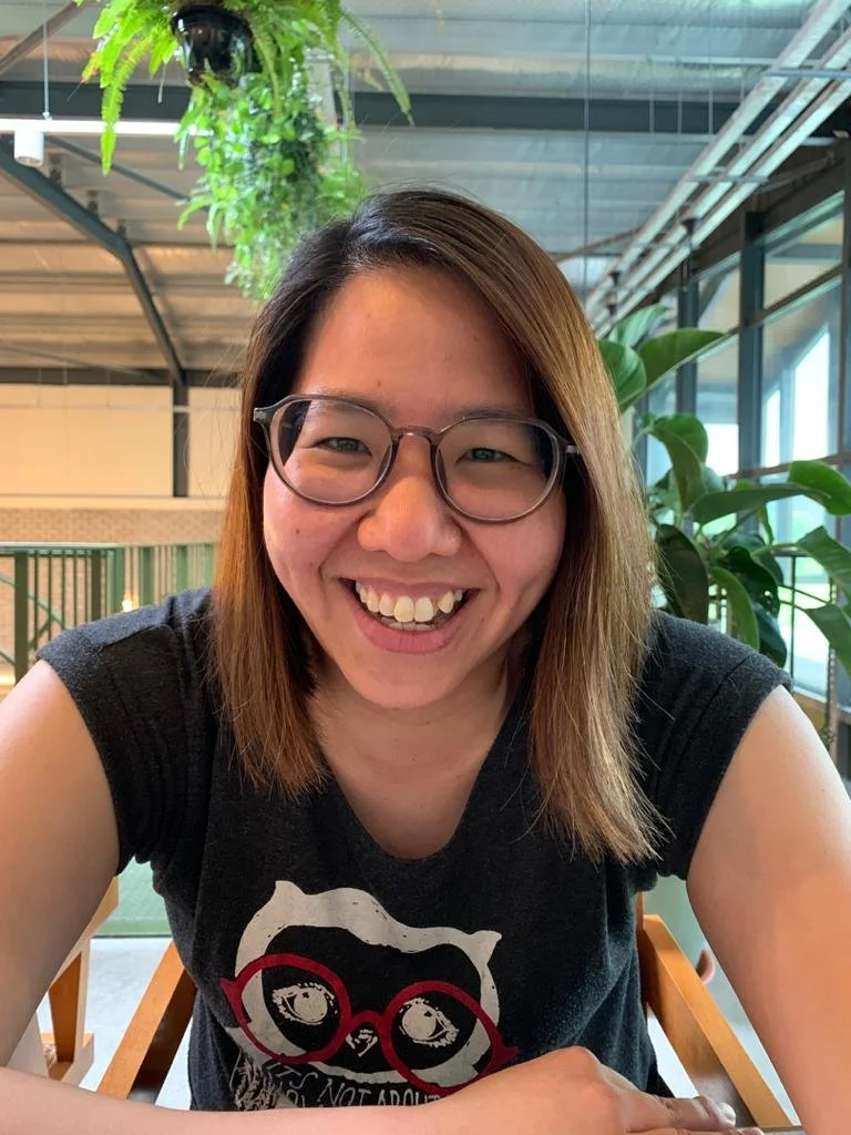 A woman with shoulder-length brown hair, wearing glasses and a black sleeveless shirt with a graphic of a dog's face and red glasses, smiling at the camera inside a modern building with large windows, green plants, and metal ceiling beams.