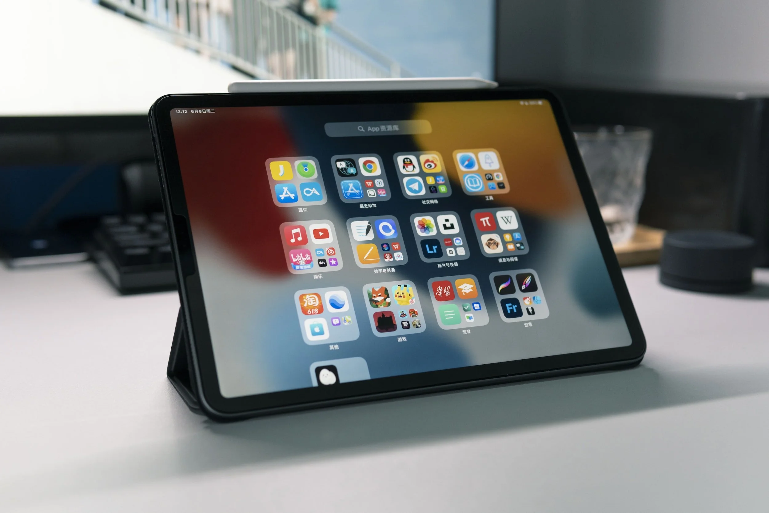 A tablet with a black case in a landscape orientation, displaying a grid of app icons on a blurred background, placed on a white desk.
