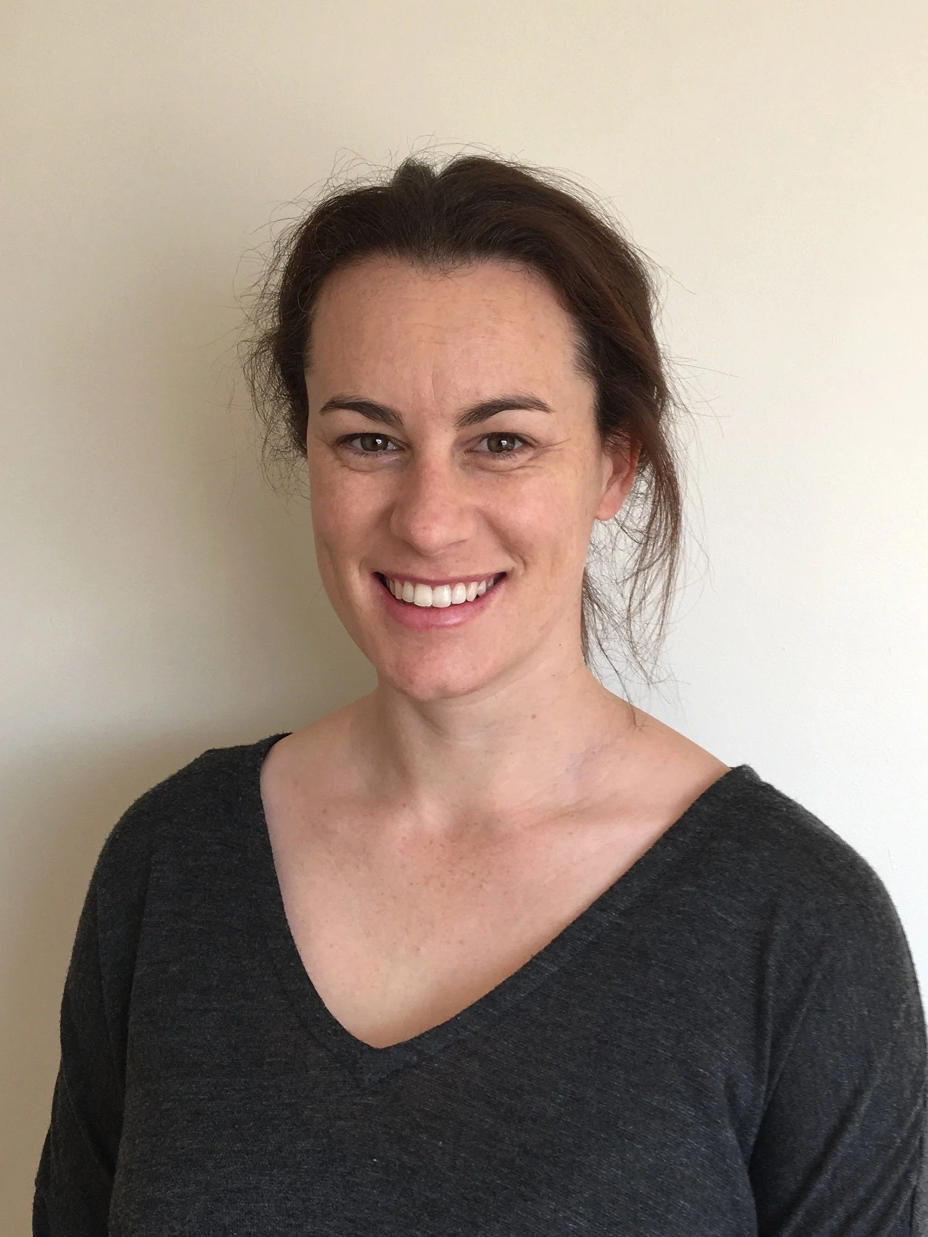A woman with dark brown hair, smiling, wearing a dark gray V-neck shirt, standing against a plain light-colored wall.