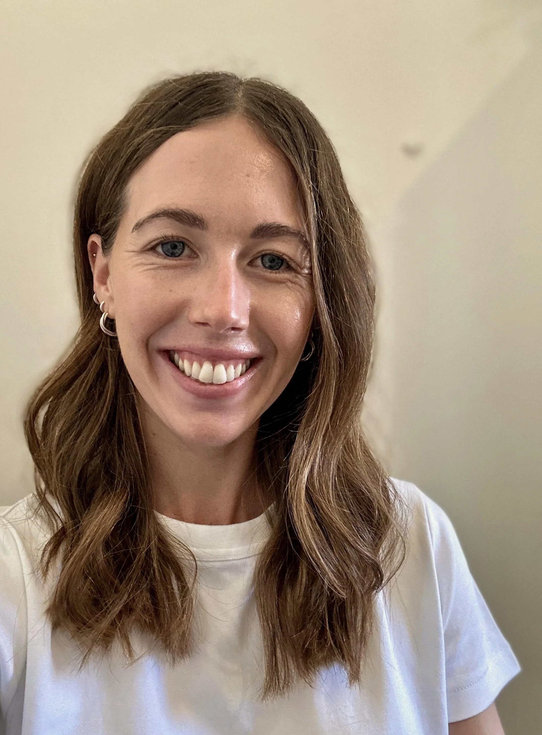 A woman with wavy brown hair, blue eyes, and multiple earrings in her left ear, smiling while wearing a white t-shirt, against a plain beige wall.