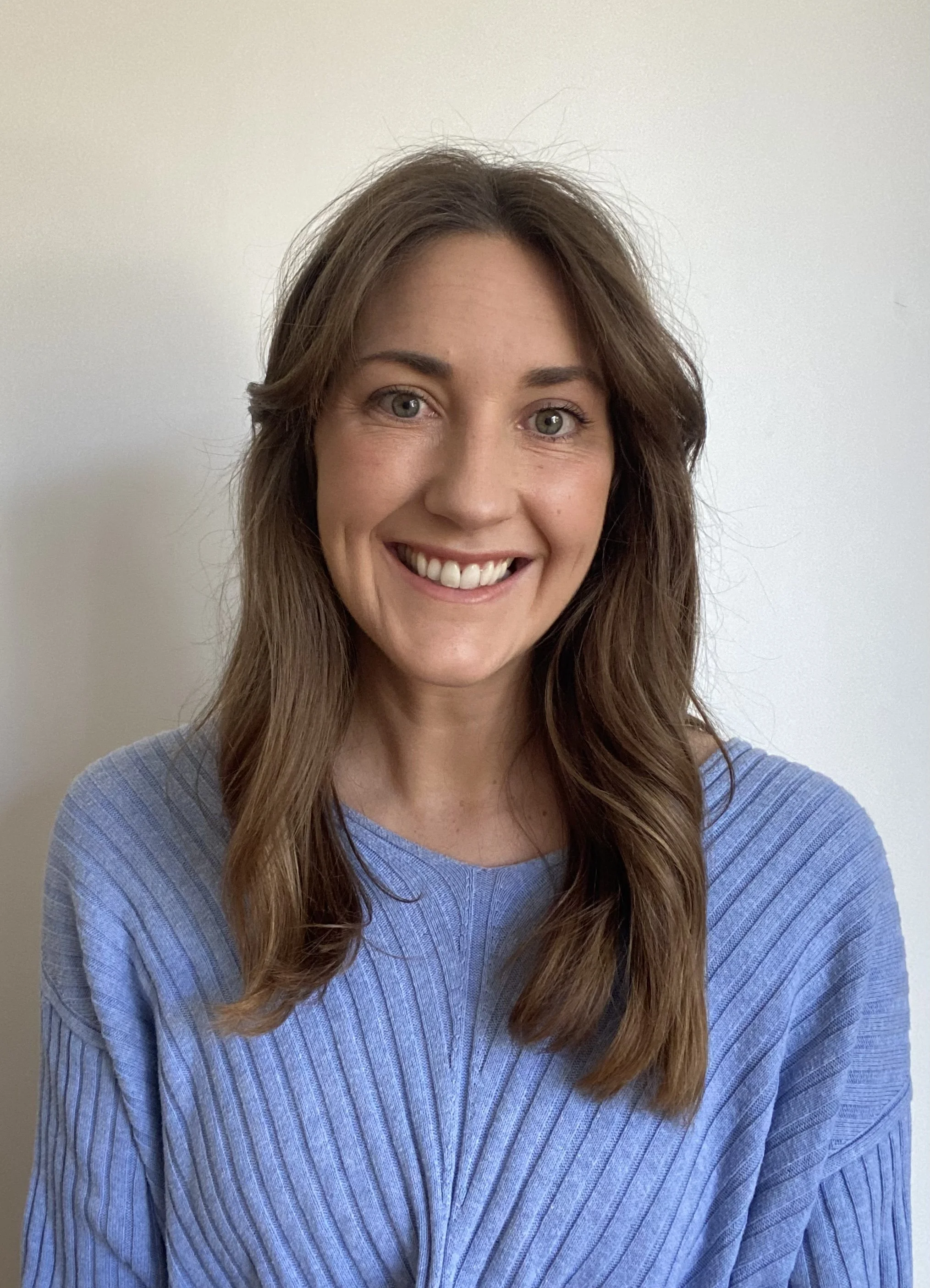 A woman with long wavy brown hair and blue eyes smiling, wearing a light blue top, standing against a plain white wall.