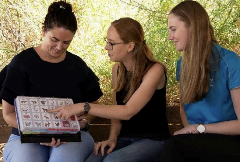 Three women sitting outdoors looking at a book or folder with images and text.