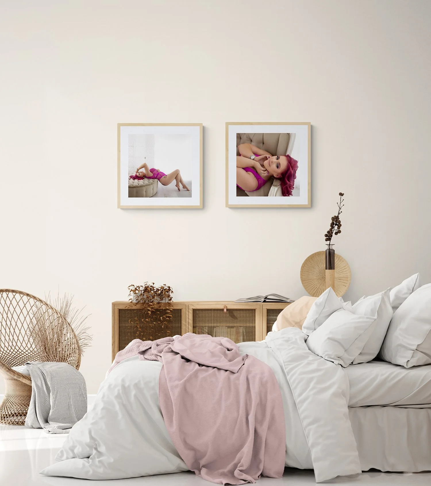 A bedroom with a white bed, pink blanket, and two framed photos of a woman with pink hair, one lying on a couch and the other reclining, on the wall above the bed. There is a rattan chair with a gray blanket on the left and a wooden sideboard with plants and a vase on the right.