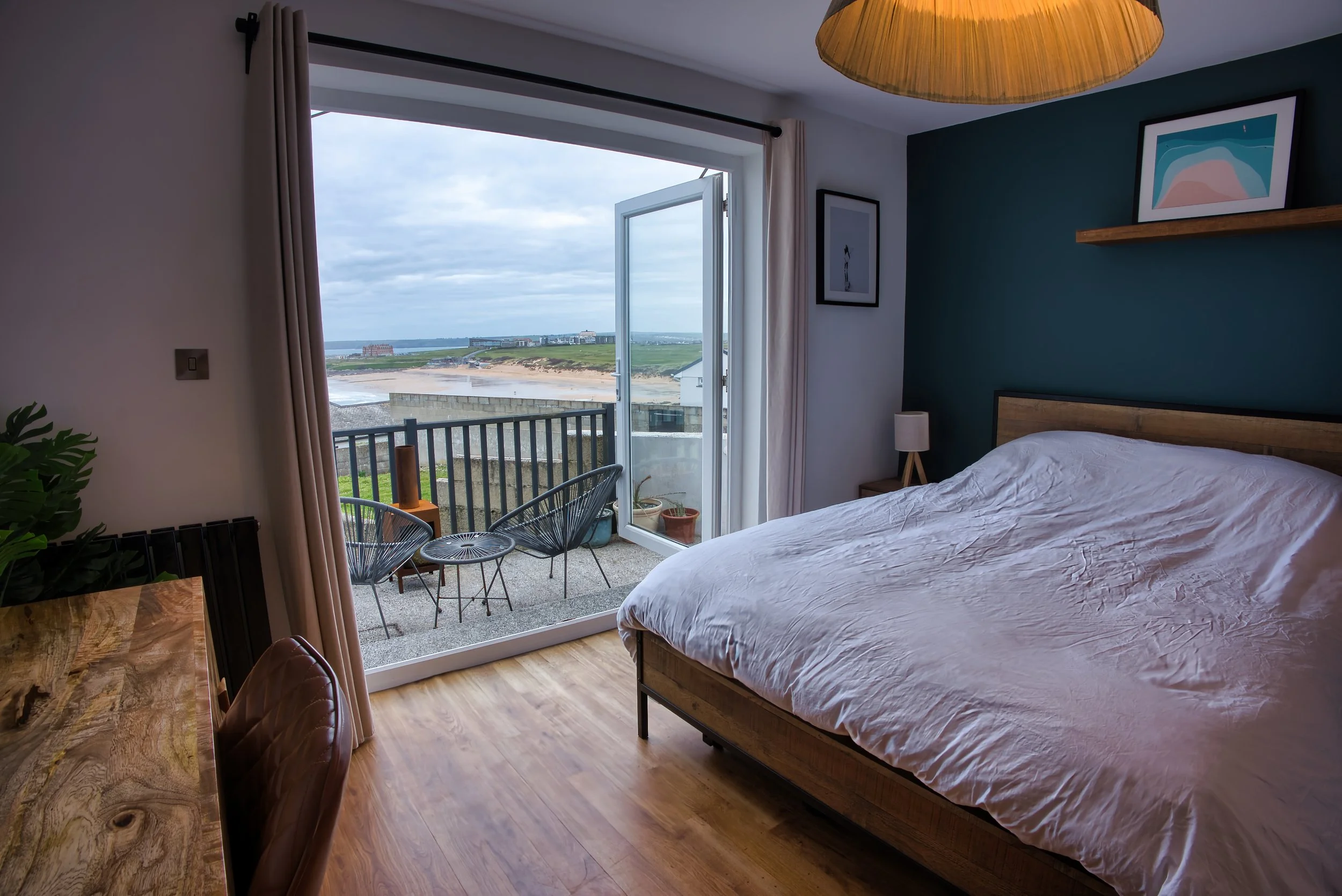 A bedroom with a bed, a nightstand and a lamp, open balcony door showing a view of a beach, grassy dunes, and a cloudy sky.