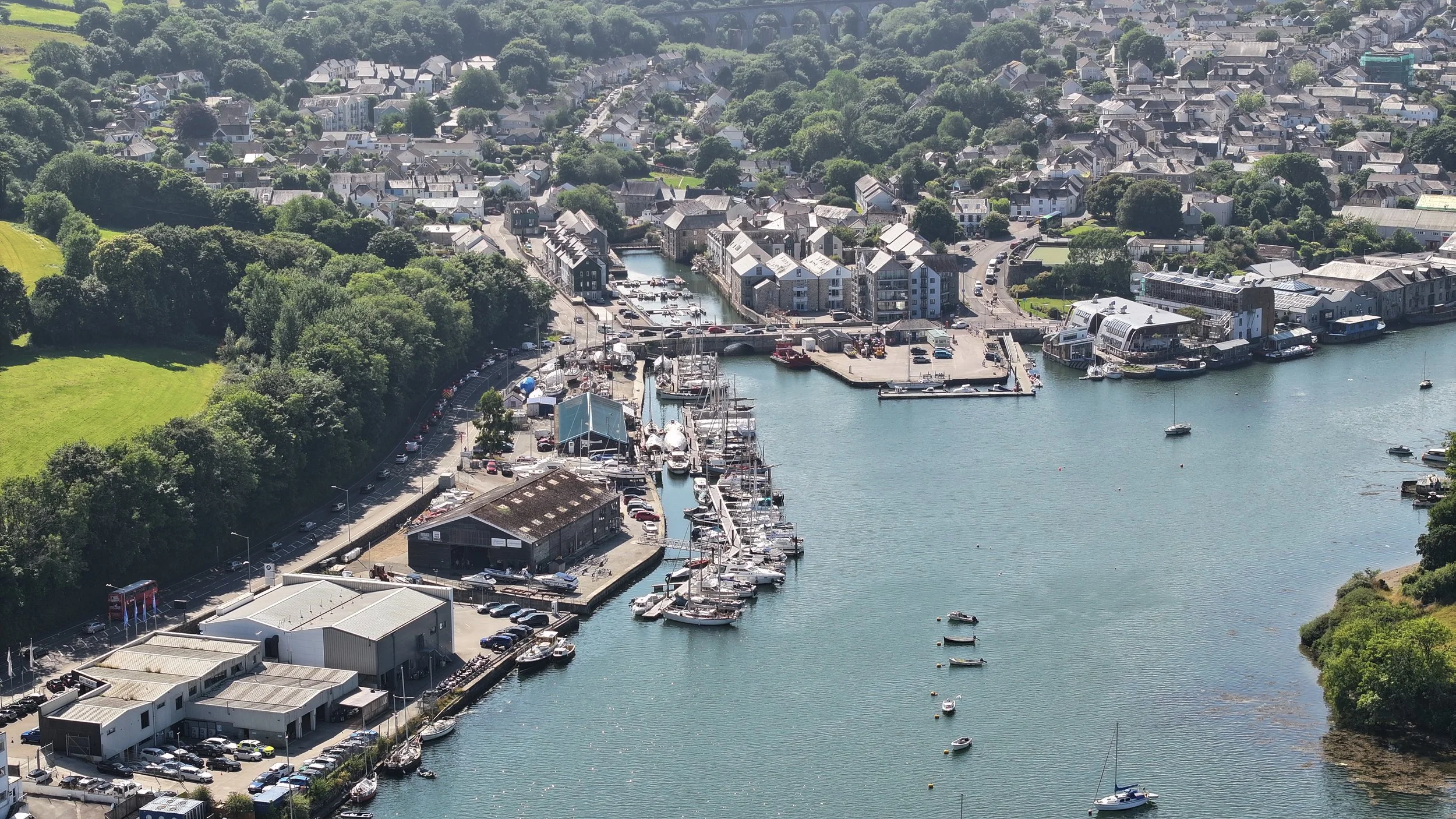 An aerial view of a harbor with boats docked, surrounded by buildings, greenery, and a residential area in the background.