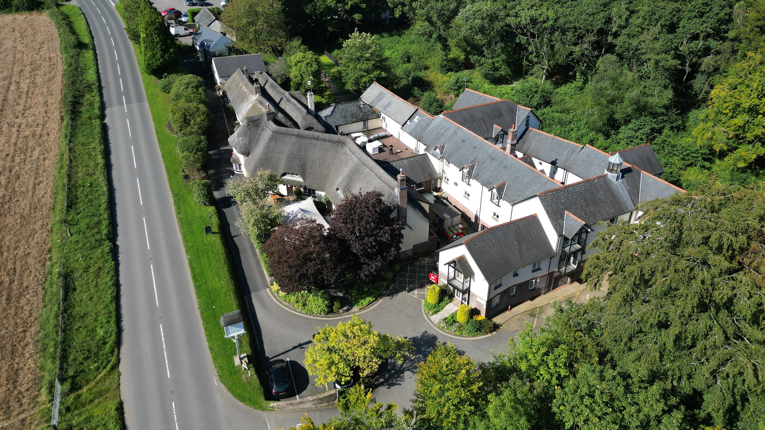 An aerial view of a large white house with a gray roof, surrounded by trees and a parking lot, next to a winding road in a rural area.