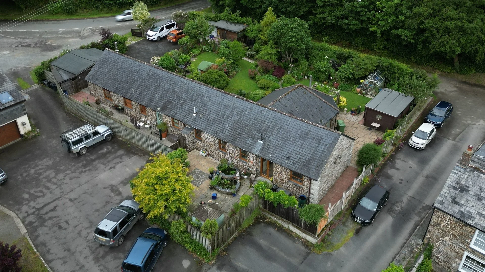 Aerial view of a stone house with a garden, surrounded by parked cars on a paved lot, with trees and other houses nearby.