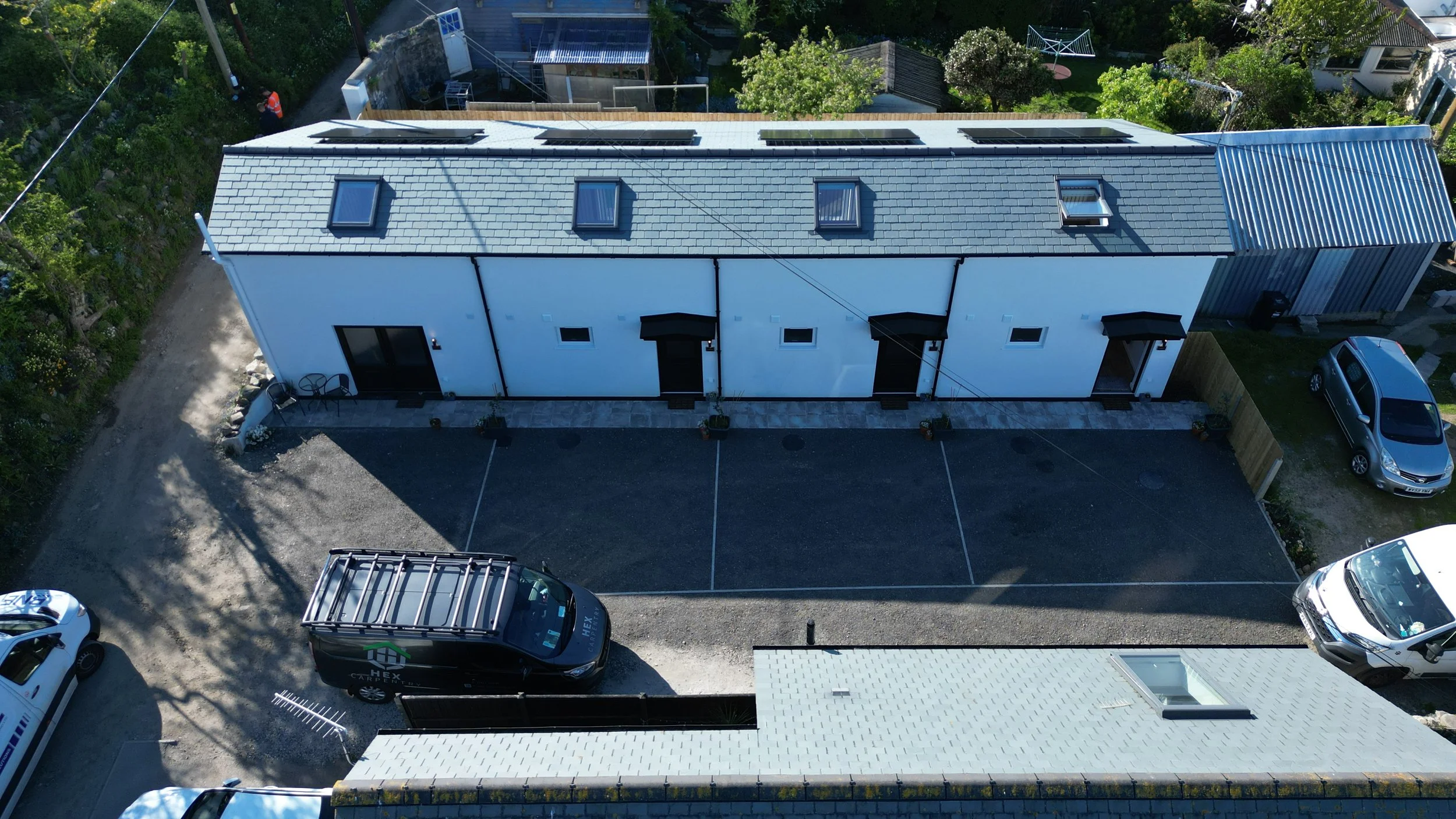 An aerial view of a modern two-story white house with a dark roof that has multiple skylights and a small balcony. There are parked cars in front and surrounding the house, with trees and neighboring houses in the background.