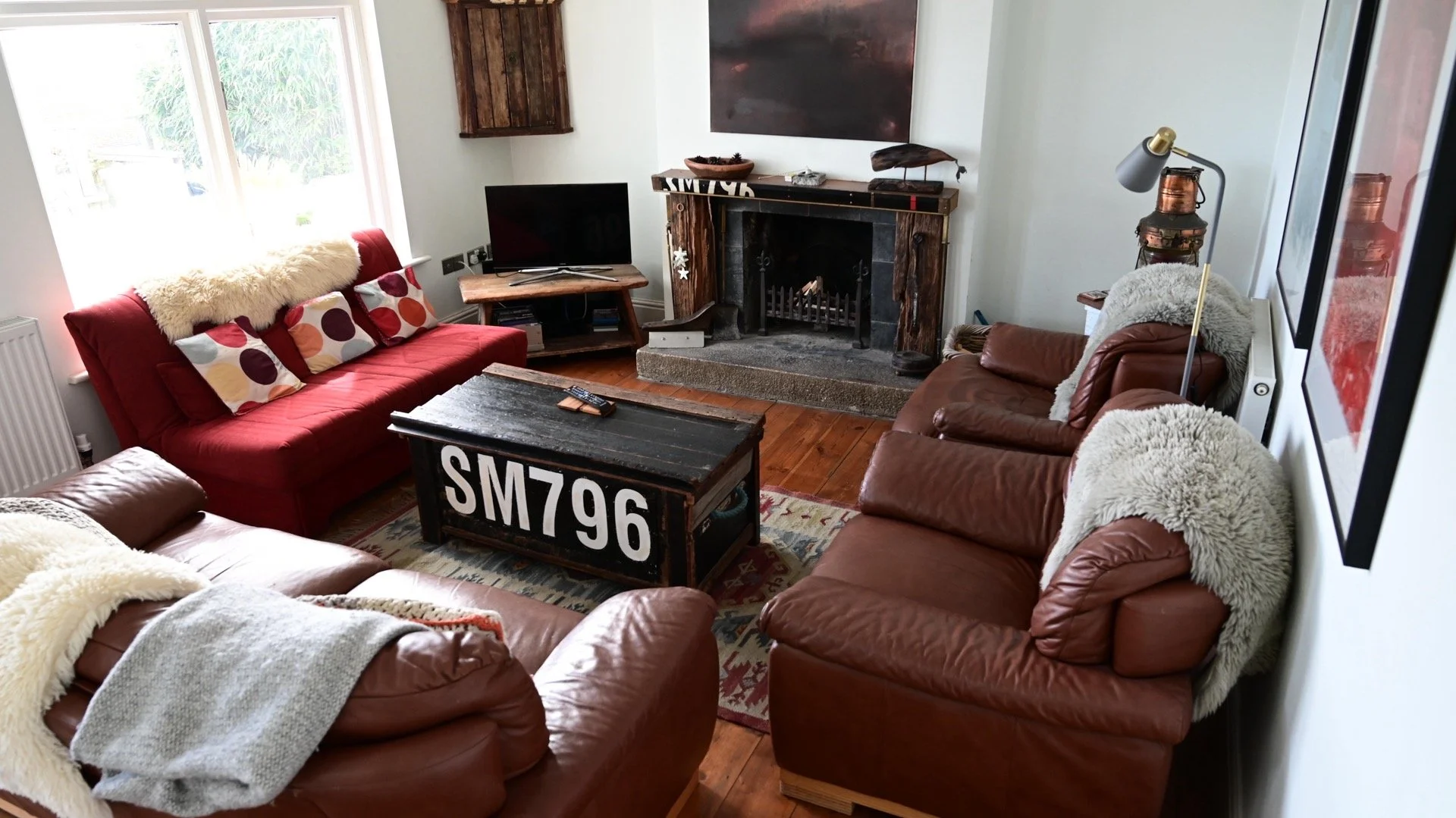 A cozy living room with two brown leather sofas and a red fabric sofa, all adorned with cream, sherry, and patterned throw pillows. A rustic wooden coffee table with white painted letters and numbers sits in the center. There is a fireplace with a wooden surround and a TV on a small wooden stand to the left of the fireplace. Large windows let in natural light, and there are artwork and decor on the walls and fireplace mantel. The floor is hardwood, and a patterned area rug partially covers it.