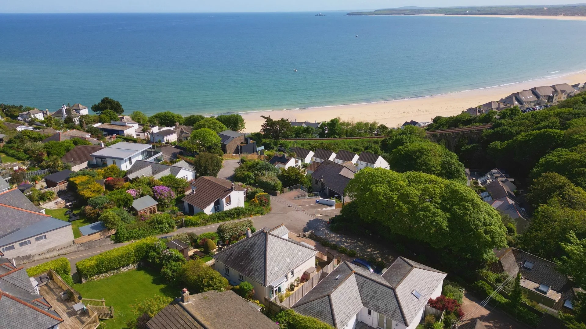 Aerial view of a coastal residential neighborhood with houses surrounded by green trees, overlooking a sandy beach and ocean with boats and distant land on the horizon.