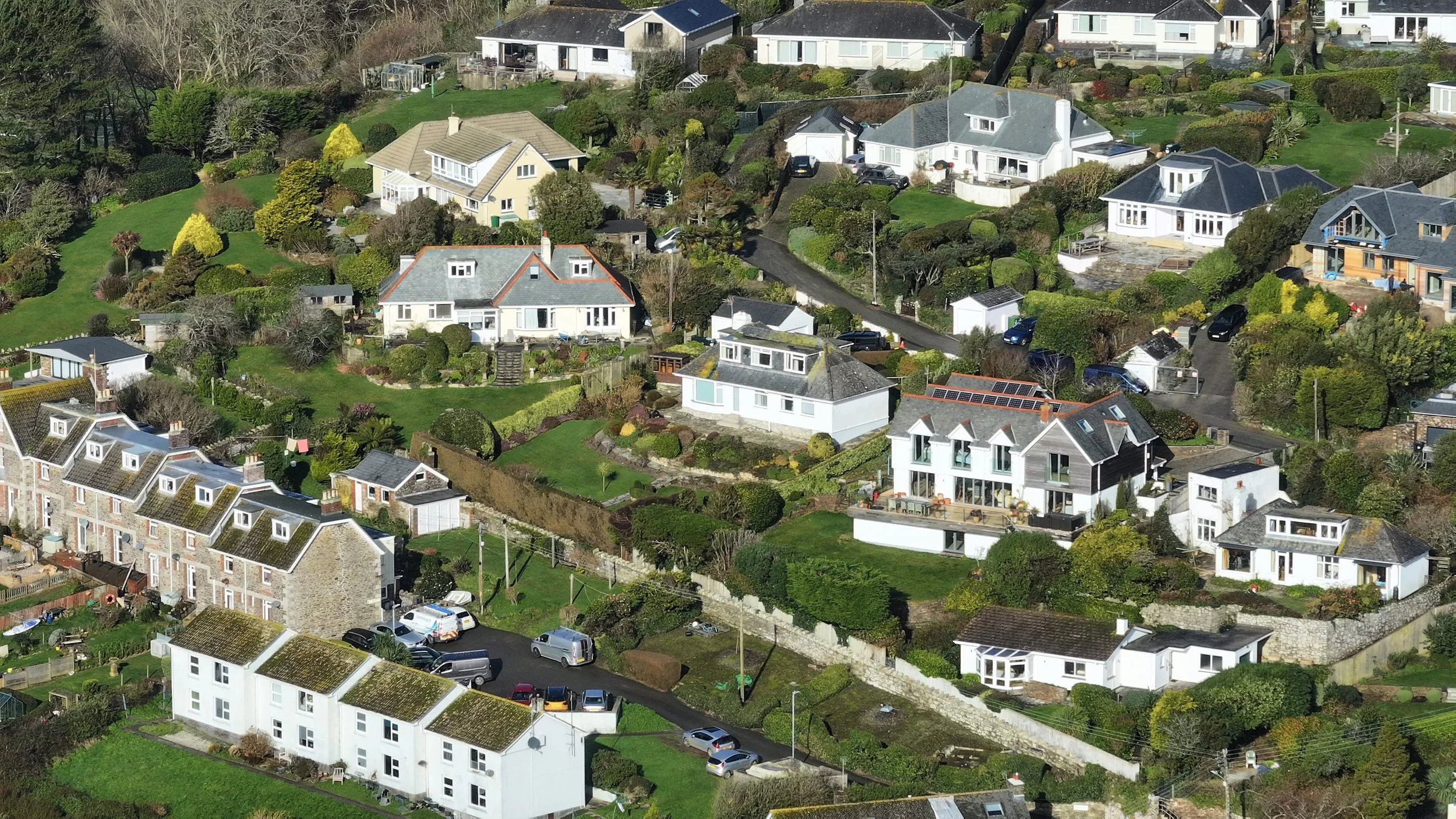 A hilly residential neighborhood in St Mawes, Cornwall with a variety of houses, including some with solar panels on the roofs. The area is lush with green lawns, trees, shrubs, and well-maintained gardens.