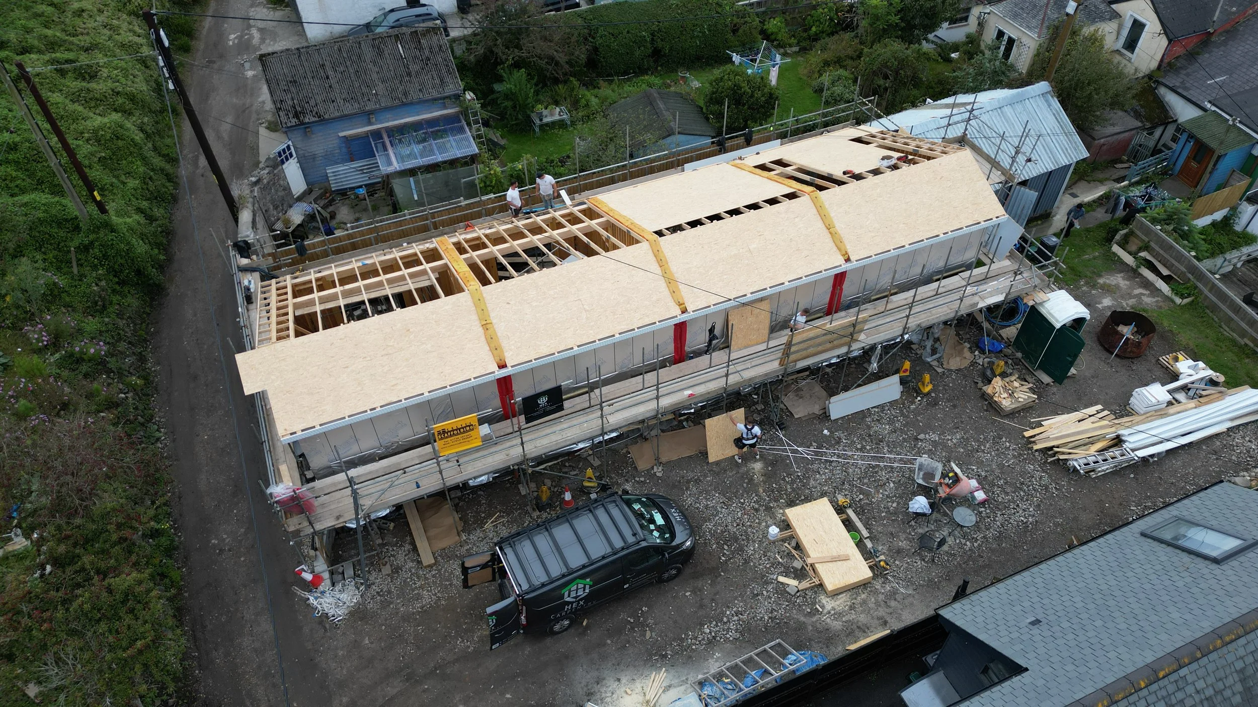 An aerial view of a construction site where a building is being built with wooden framing. Several workers are on site, and various construction materials and equipment are scattered around.