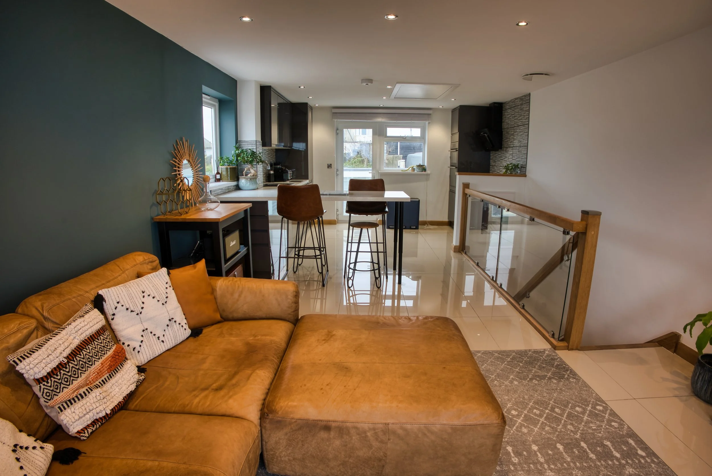 Modern living room with a tan leather sofa decorated with patterned pillows, a wooden side table, and decorative items. The room features a white ceiling with recessed lighting, a green accent wall, and large windows leading to an outdoor area. Part 