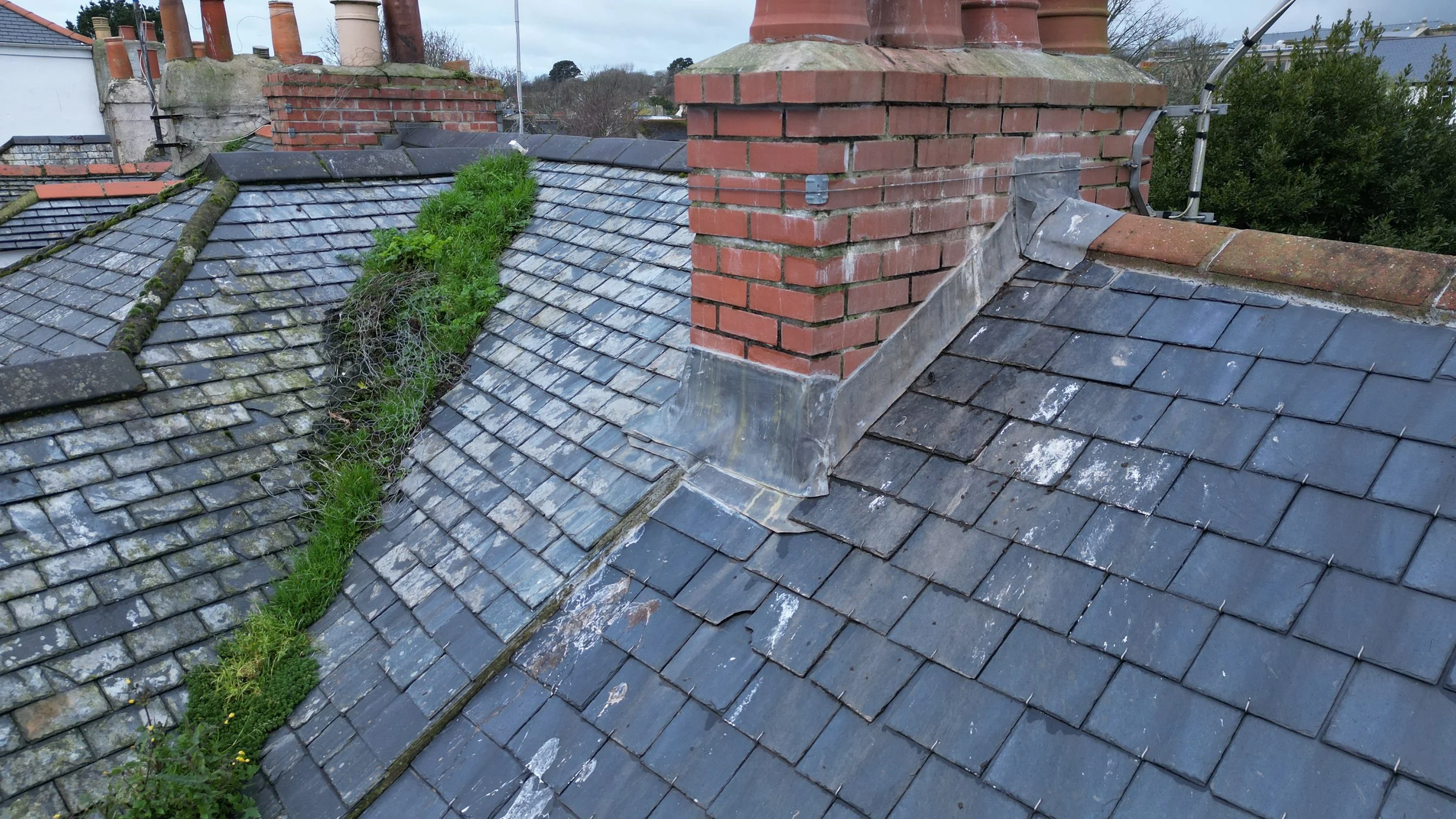The image shows a rooftop with old, weathered gray slate tiles. There is a large brick chimney with multiple pipes protruding from it, some covered with metal flashing. Green moss and plants are growing in the seams between the tiles, especially alon