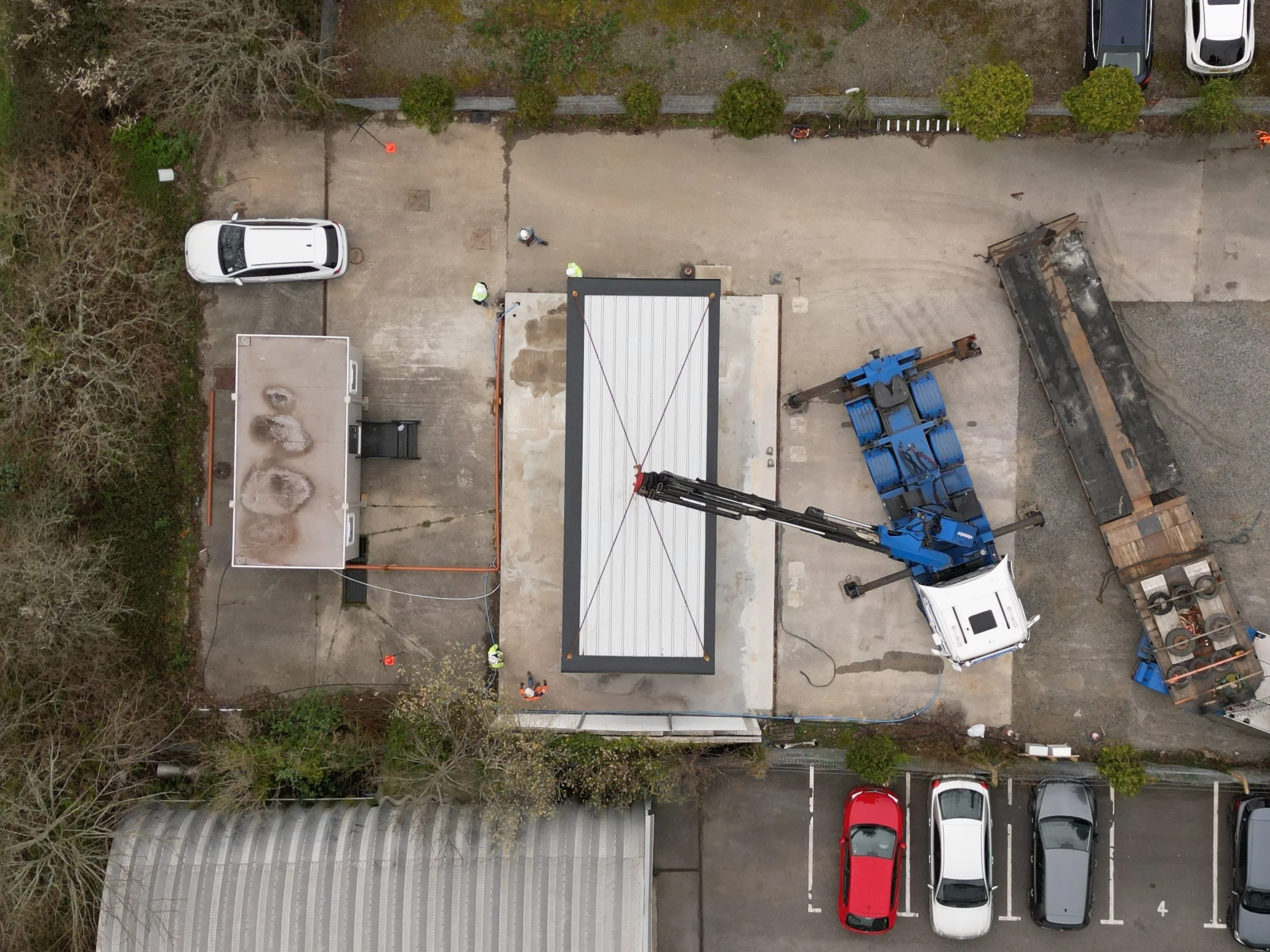 An aerial view of a construction site with several workers, construction equipment, a partially assembled roof, parked cars, and trees surrounding the area.