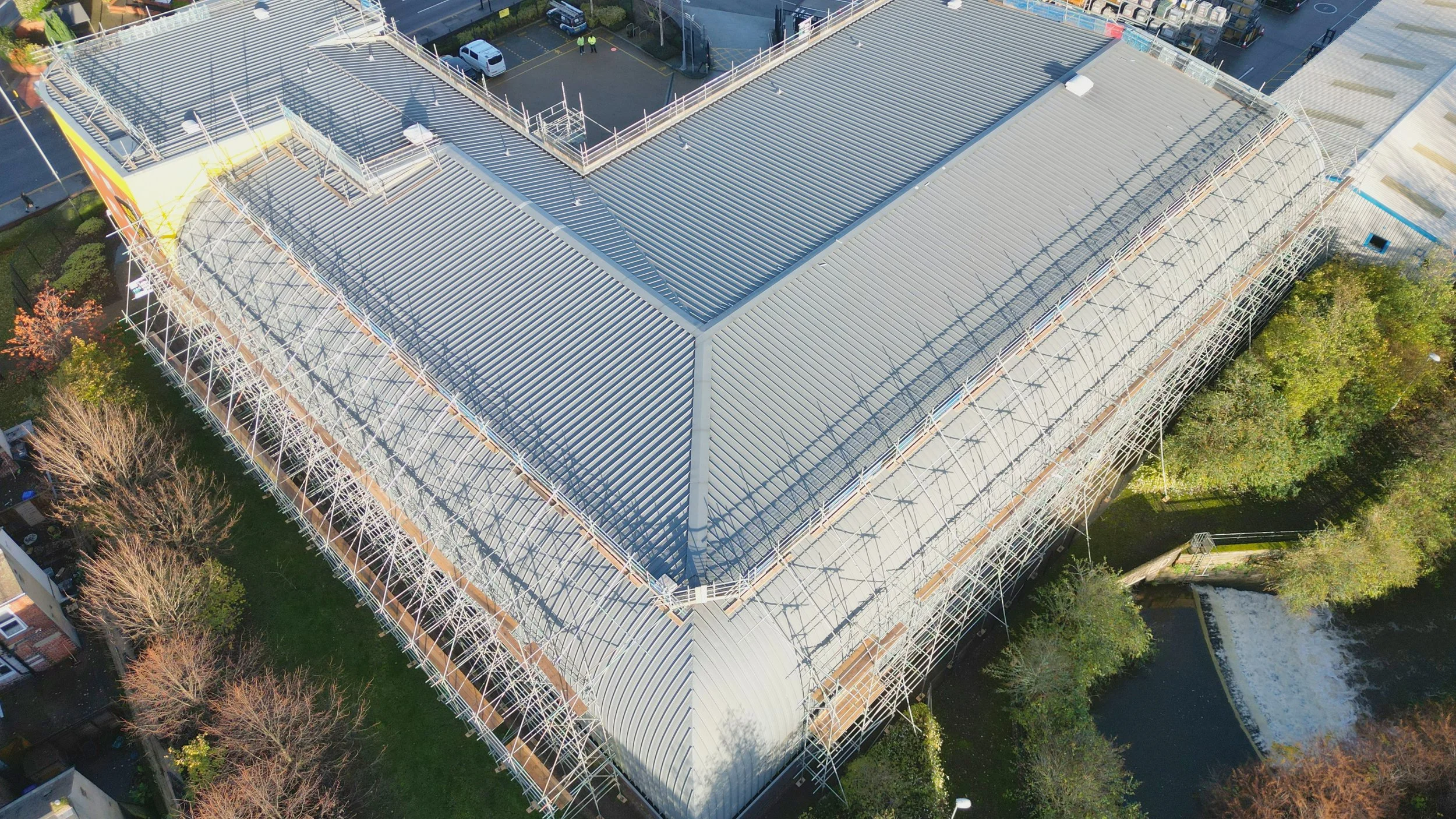 Aerial view of a building under construction with steel roofing and scaffolding, surrounded by trees and parking lots.