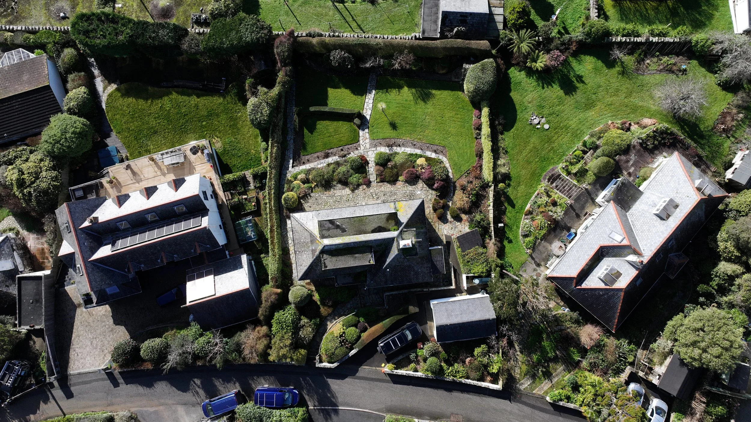 Aerial view of multiple houses with large gardens, green lawns, pathways, trees, and shrubs along a curved street.