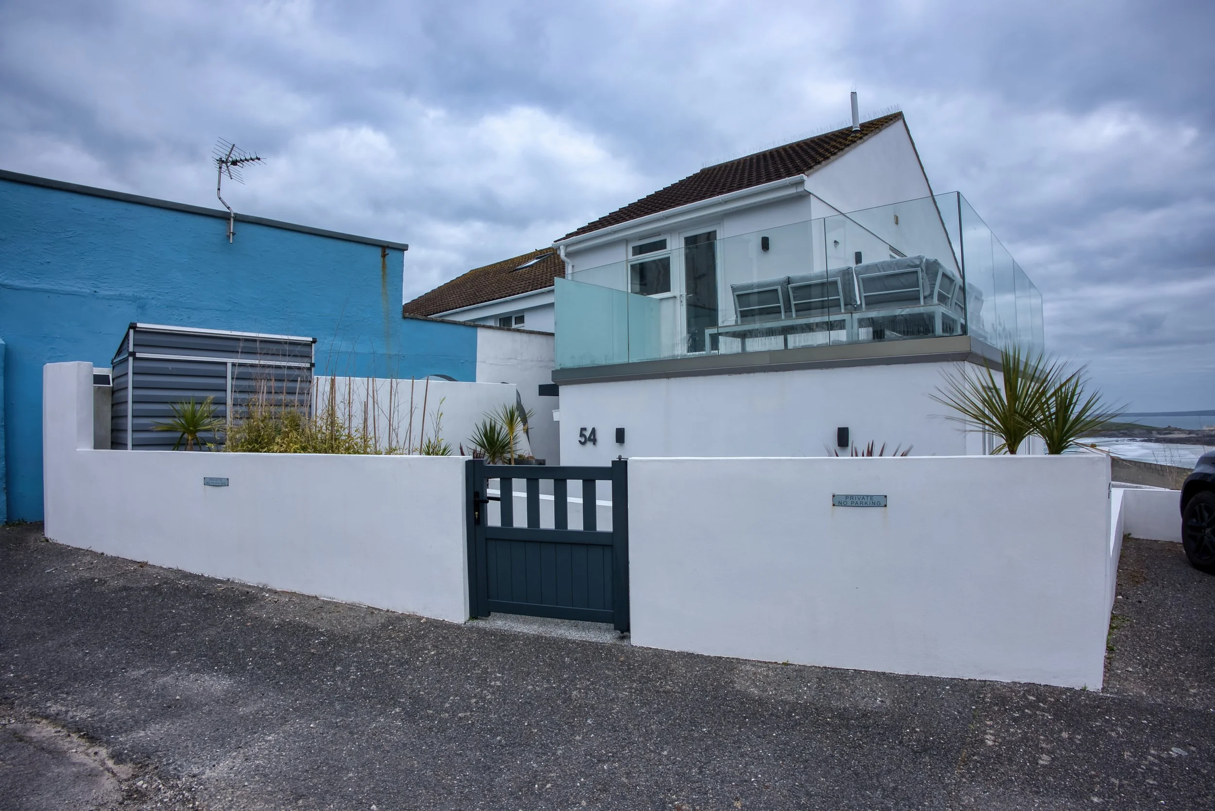 A modern house with white walls and a brown tiled roof, featuring a glass balcony and multiple windows. The house is surrounded by a white wall with the number 54 and a gate labeled 'Private No Parking.' There is a blue building to the left with an a