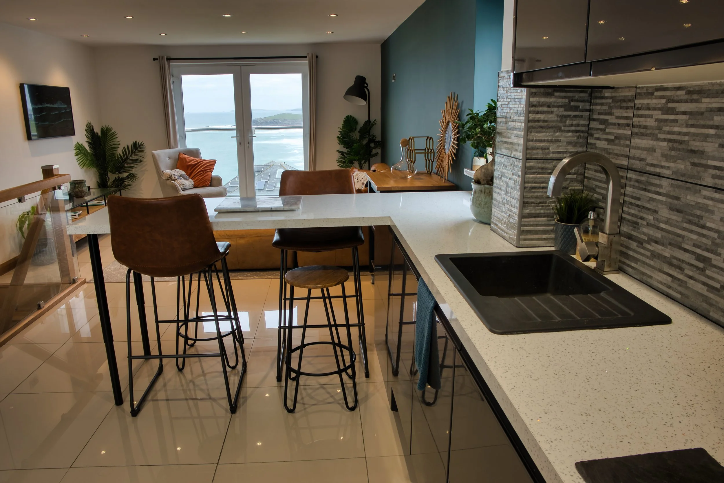 Modern open-concept living space with a kitchen island, bar stools, a cozy seating area with a chair and orange pillows, large windows with a view of water and hills, and decorative plants and artwork.