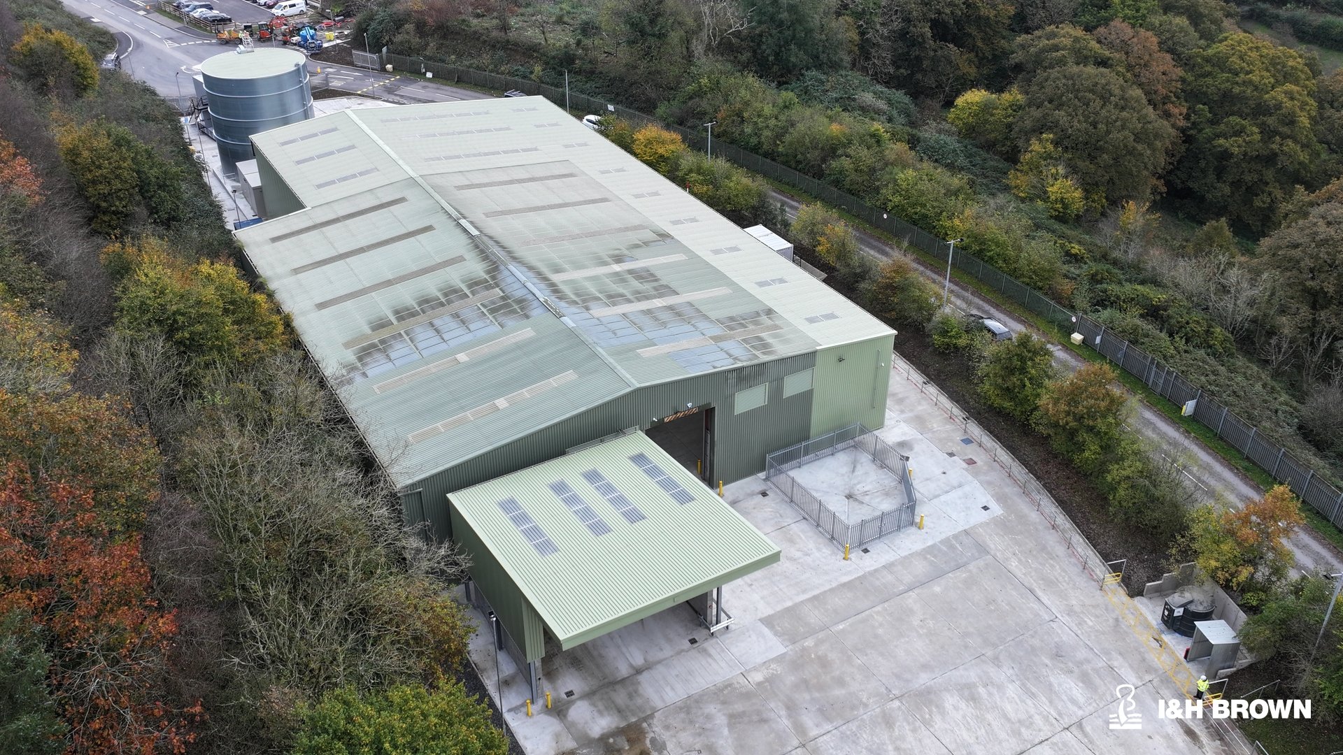 Aerial view of a large industrial building with a light green roof and solar panels, surrounded by trees and a parking lot, with a pathway and fencing nearby.