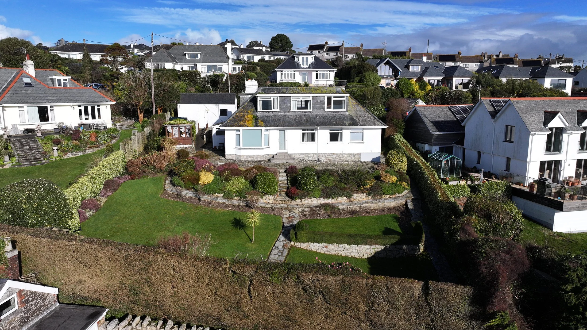 Aerial view of a residential neighborhood with white houses, well-maintained gardens, and blue sky with some clouds.