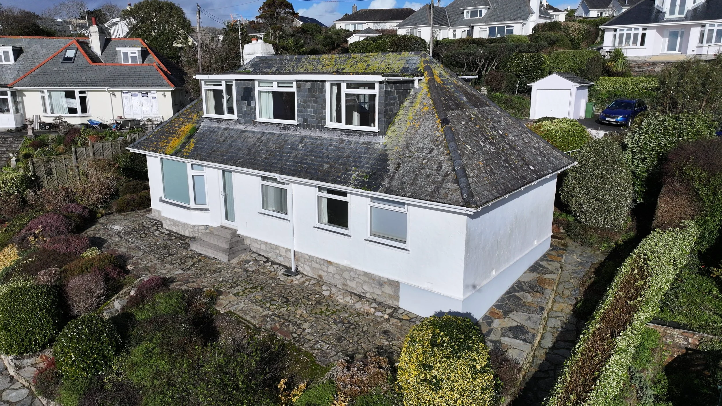 A two-story white house with a stone pathway patio, multiple windows, and a sloped mossy roof, surrounded by garden shrubs and neighbouring houses.