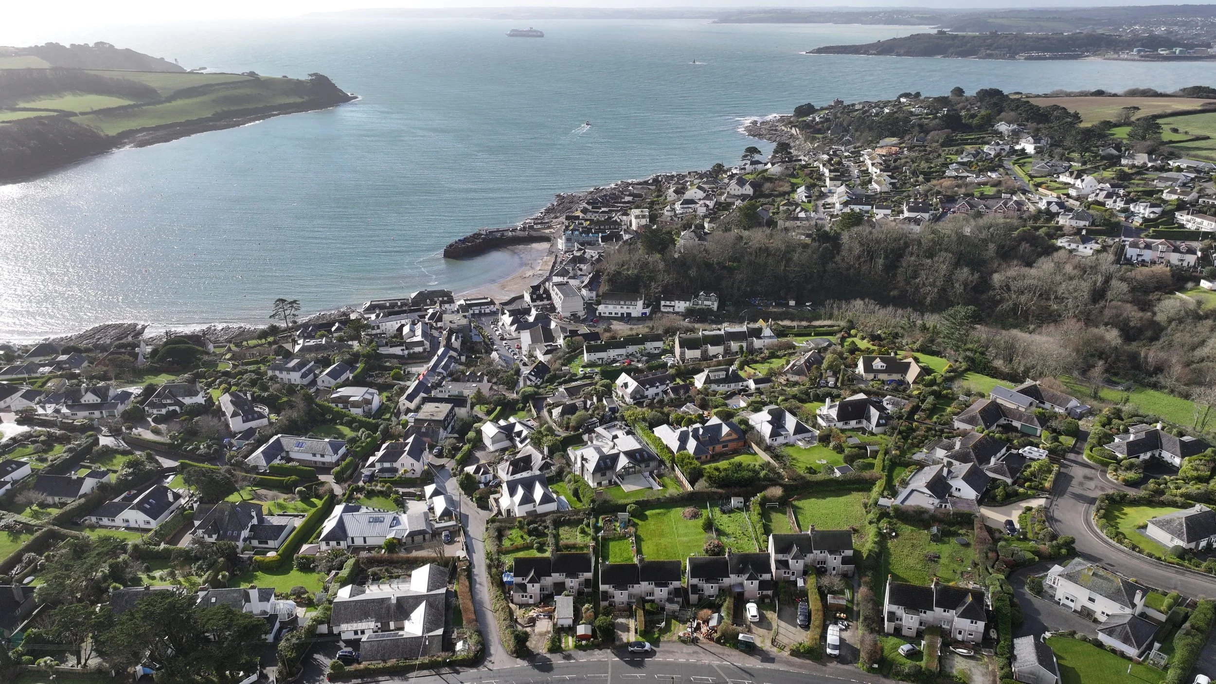 Aerial view of St Mawes in Cornwall with numerous houses, winding roads, and green gardens, next to a bay with boats and a distant shoreline.