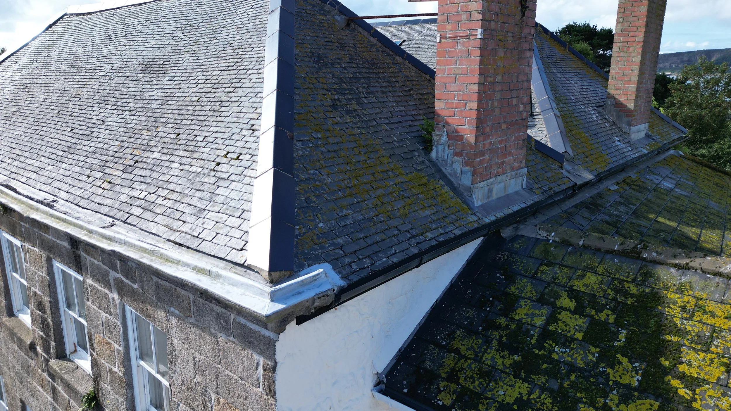 Aerial view of a building's roof showing moss and weathering on slate shingles, with brick chimneys and surrounding trees.