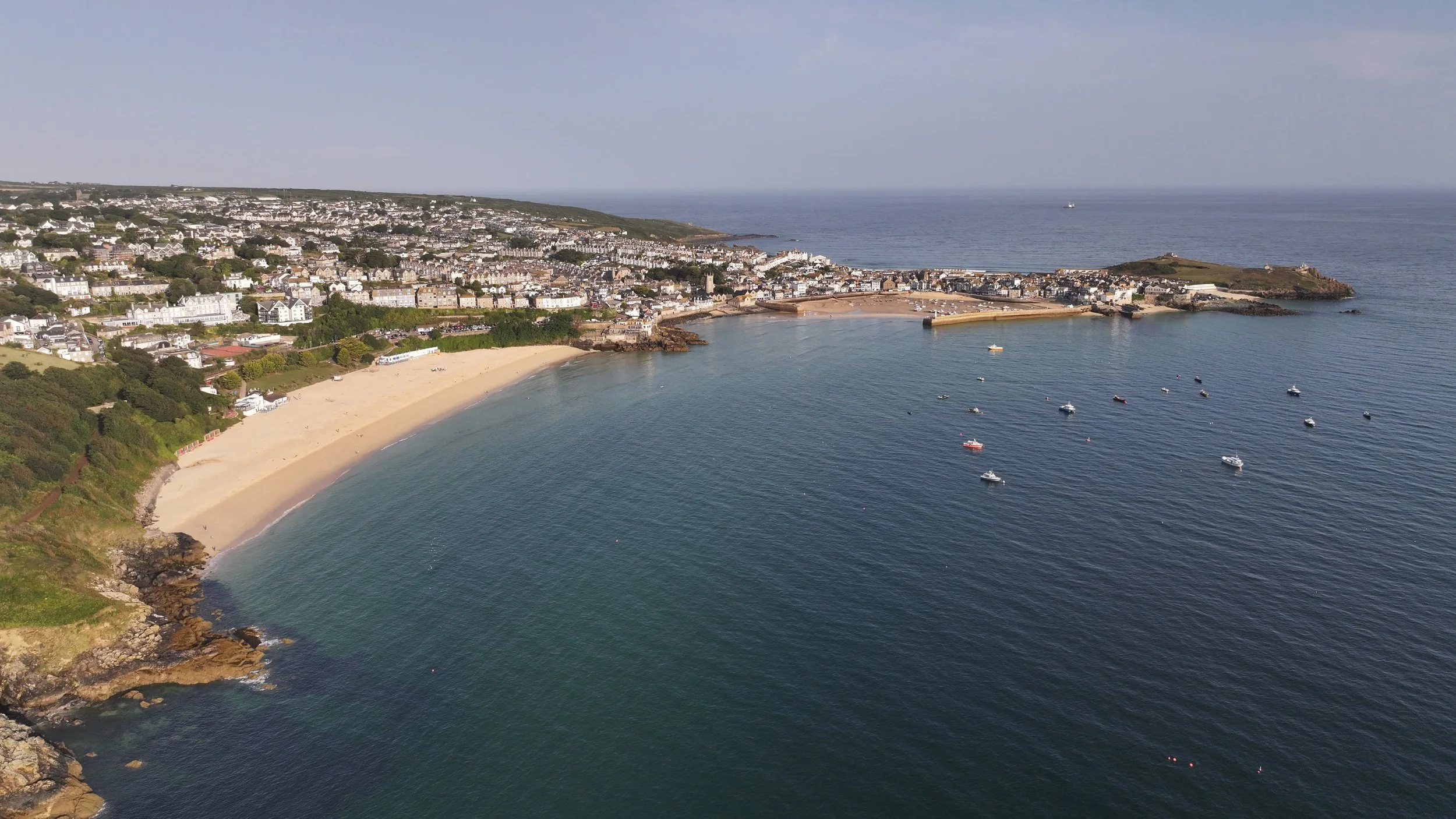 Aerial view of a coastal town with a sandy beach, boats in the water, residential buildings, and a harbor, with a hill and the ocean in the background.