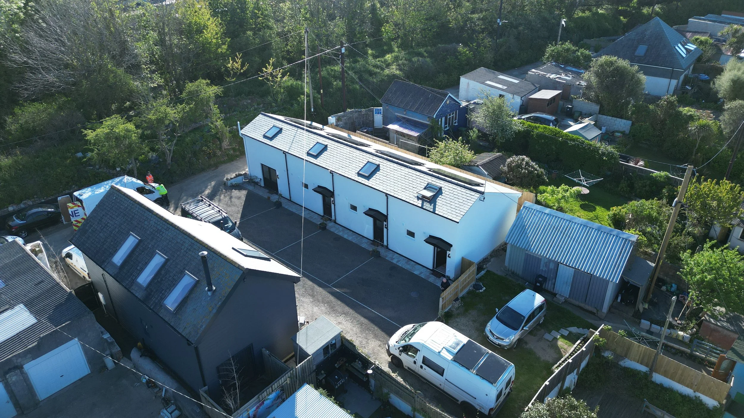An aerial view of a residential neighborhood showing various houses, cars parked on the streets, and a lush, green wooded background.