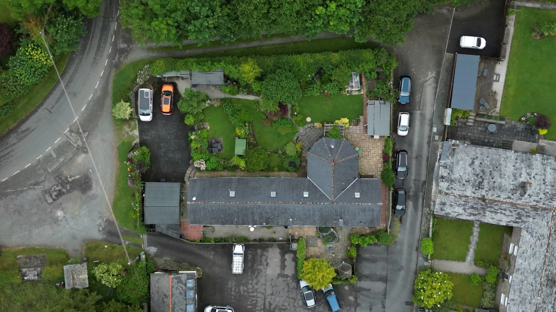 Aerial view of a garden surrounded by houses, with a driveway, cars parked, and green trees.