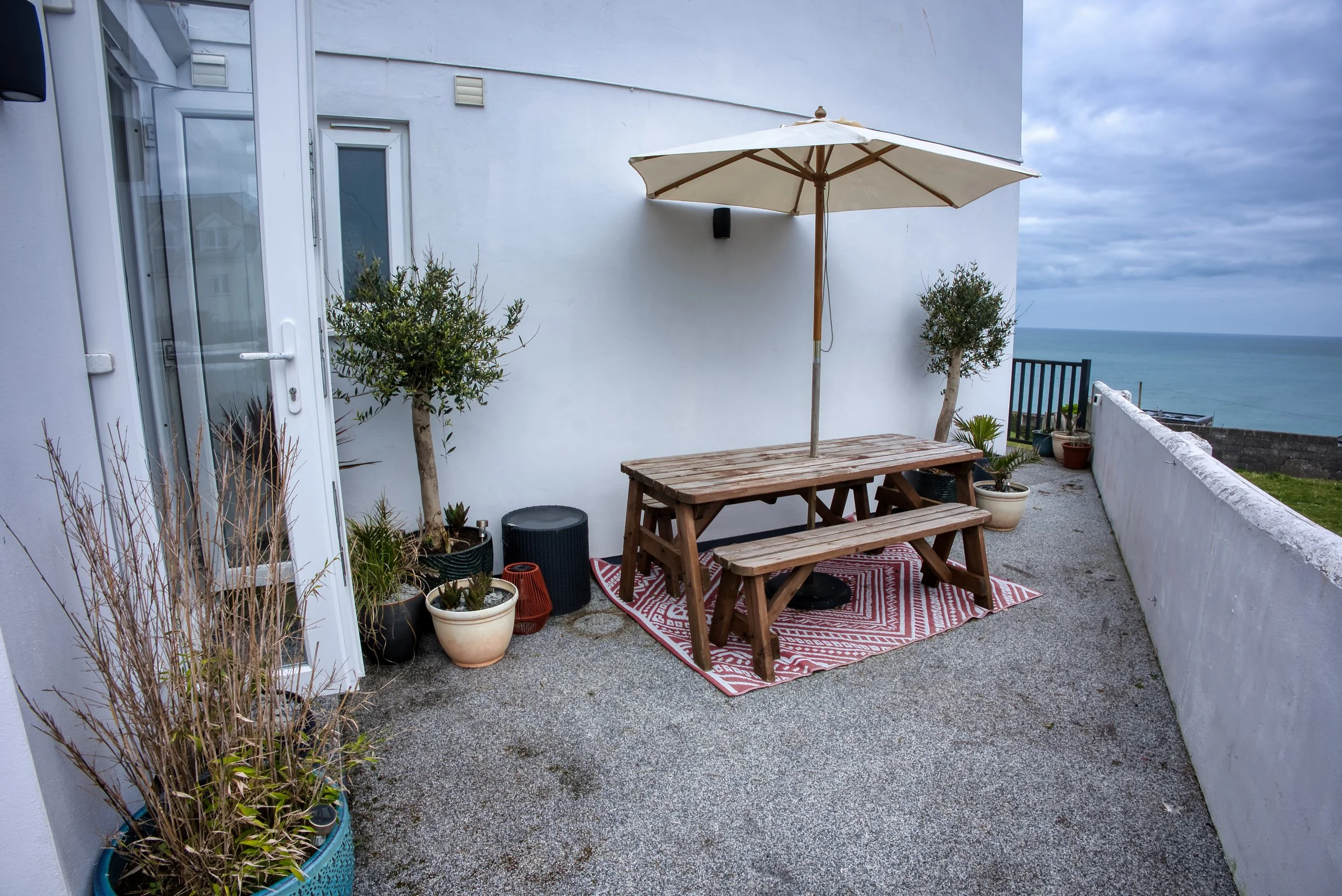 A balcony with outdoor furniture, potted plants, and an umbrella overlooking the ocean on a cloudy day.