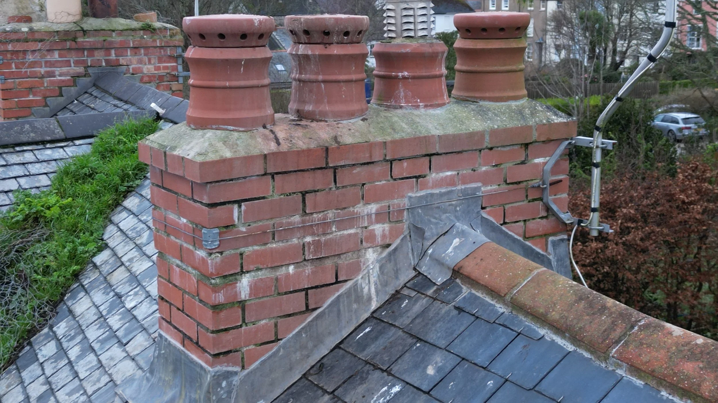 An outdoor brick chimney on a house roof with four terracotta chimney pots, some moss and weathering, and an antenna attached to the side, surrounded by trees and neighborhood houses in the background.