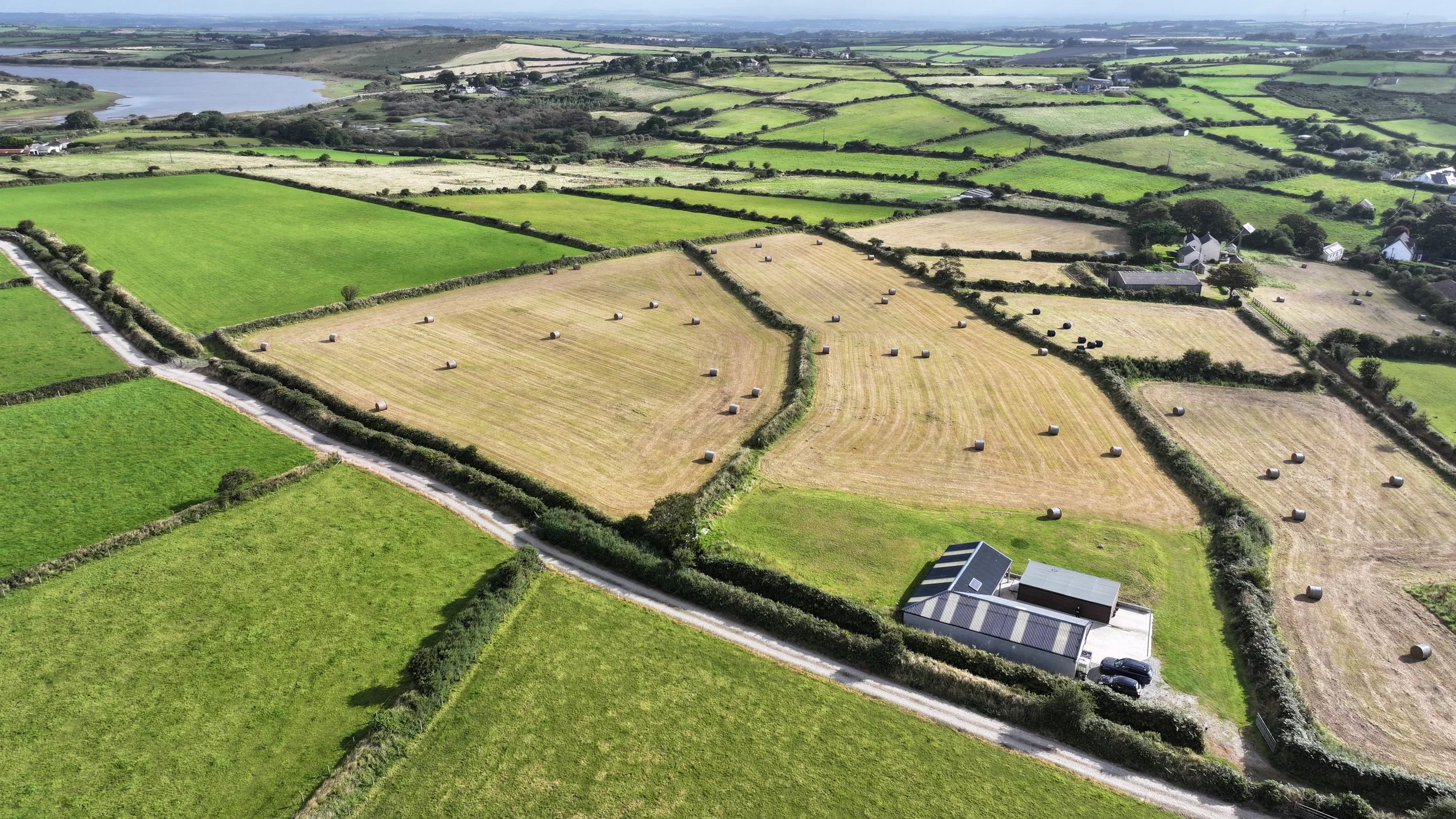 Aerial view of a patchwork of green farmland and fields with hay bales, enclosed by hedges and dirt roads near a lake in the background.