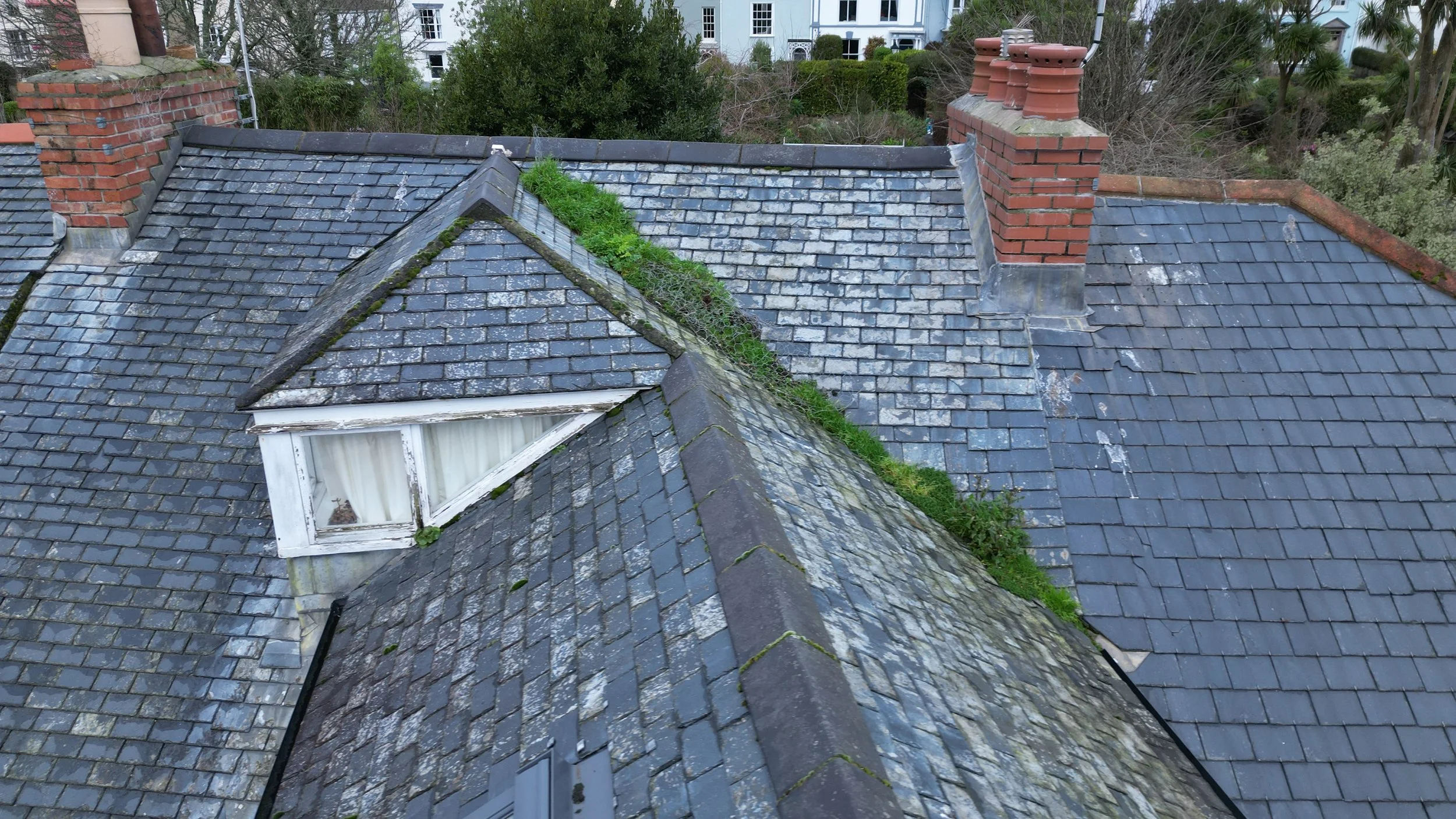 View of multiple sloped rooftops with weathered gray shingles, some covered in moss and dirt, with brick chimneys, surrounded by trees and tall buildings in the background.