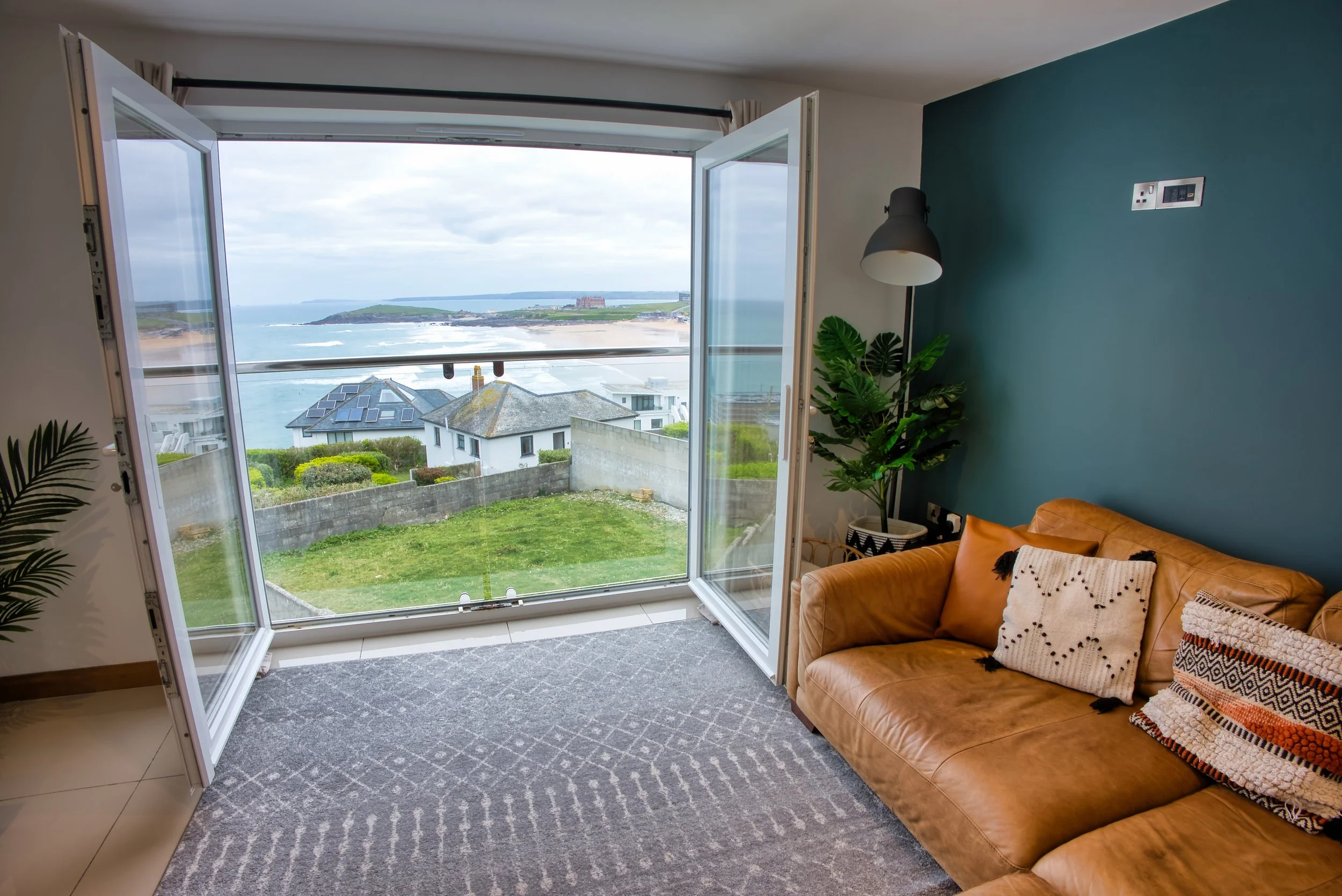 Living room with a large open window overlooking a seaside landscape, including houses, greenery, and an ocean view.