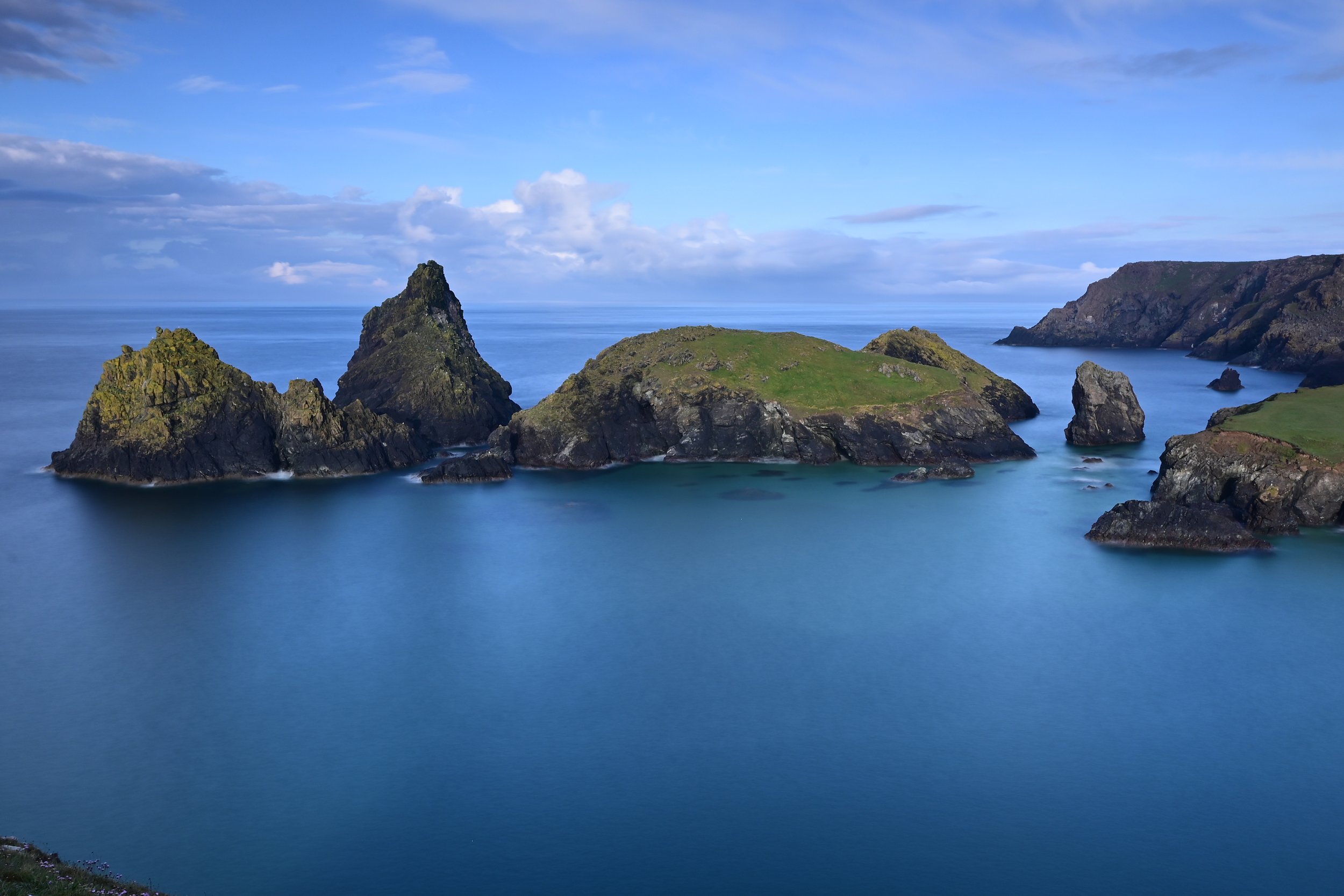 Coastal landscape with large rock formations emerging from the ocean, green grassy areas on parts of the rocks, and a partly cloudy sky.
