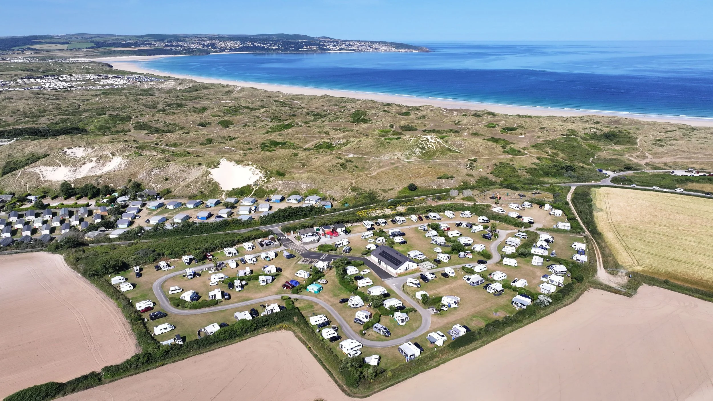 An aerial view of a coastal campground with numerous caravans and vehicles, surrounded by sandy paths, green fields, and hills overlooking a blue ocean and beach.