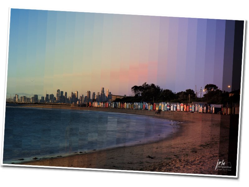 Colorful beach huts along the shoreline with a city skyline in the background at dusk or dawn, showing a sky with pastel hues.
