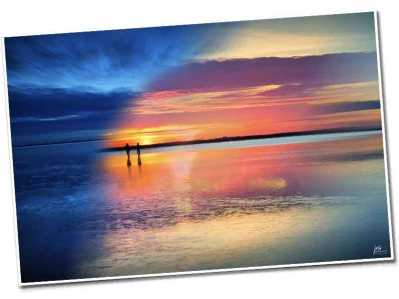 A colorful sunset over a calm body of water with two people standing near the shore.