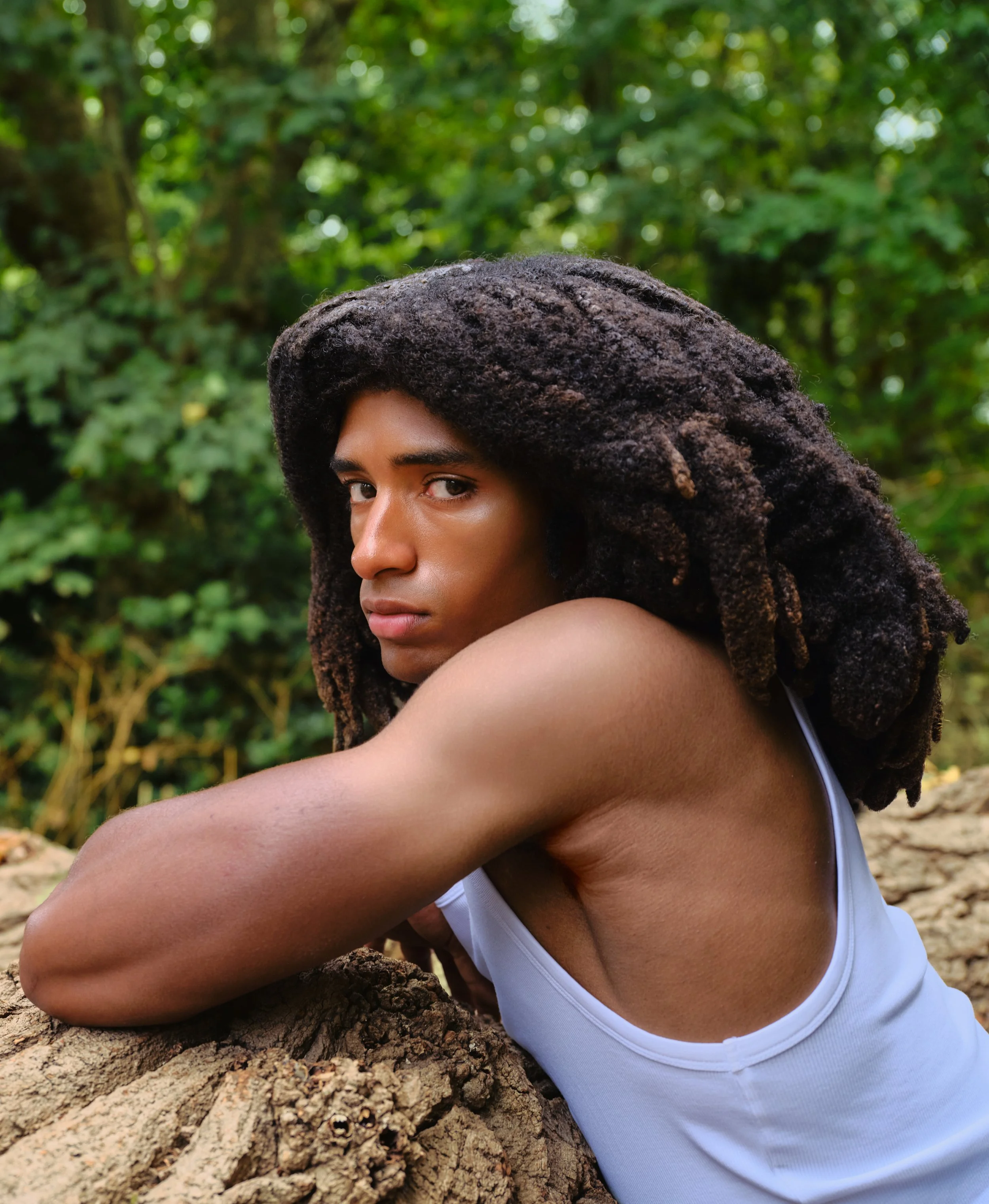 A person with dreadlocks resting on a tree root in a forest, wearing a white tank top, with a cautious expression, surrounded by green foliage.