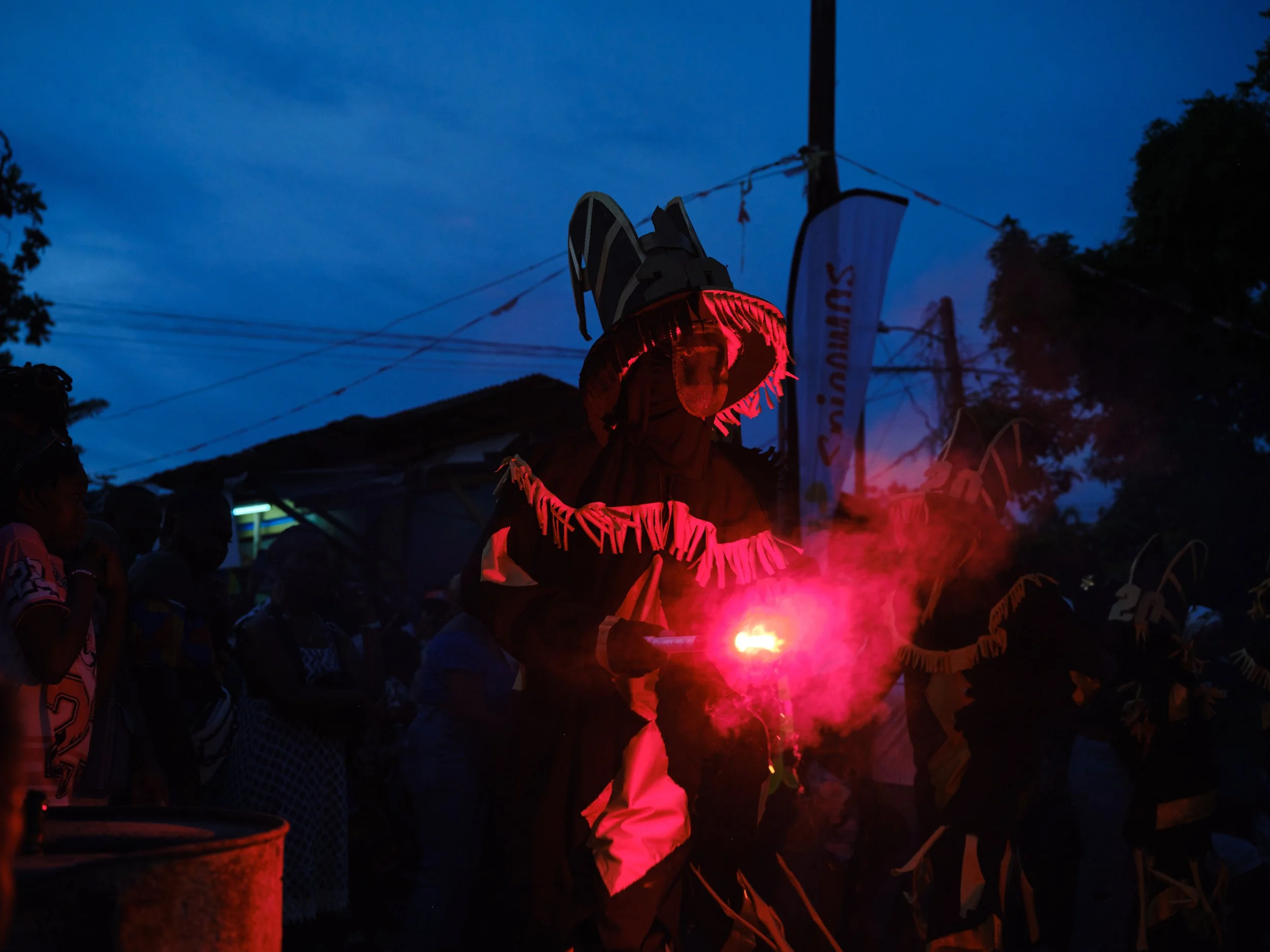 A person dressed in traditional attire holding a sparker, emitting red sparks, during a nighttime event with a crowd gathered around, and flags or banners in the background.