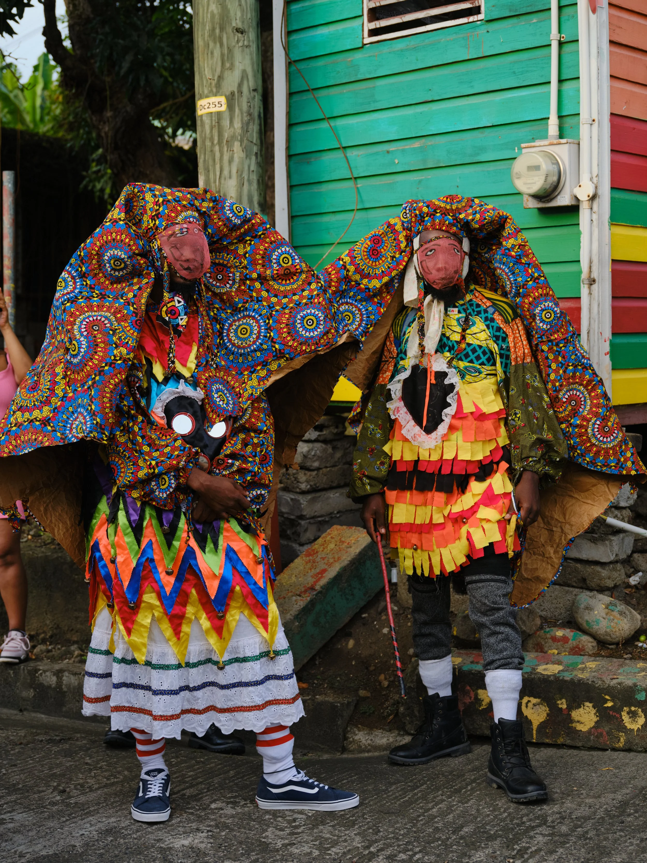 Colorfully dressed street performers in vibrant costumes and masks, with one holding a stick, standing in front of a colorful wooden building.