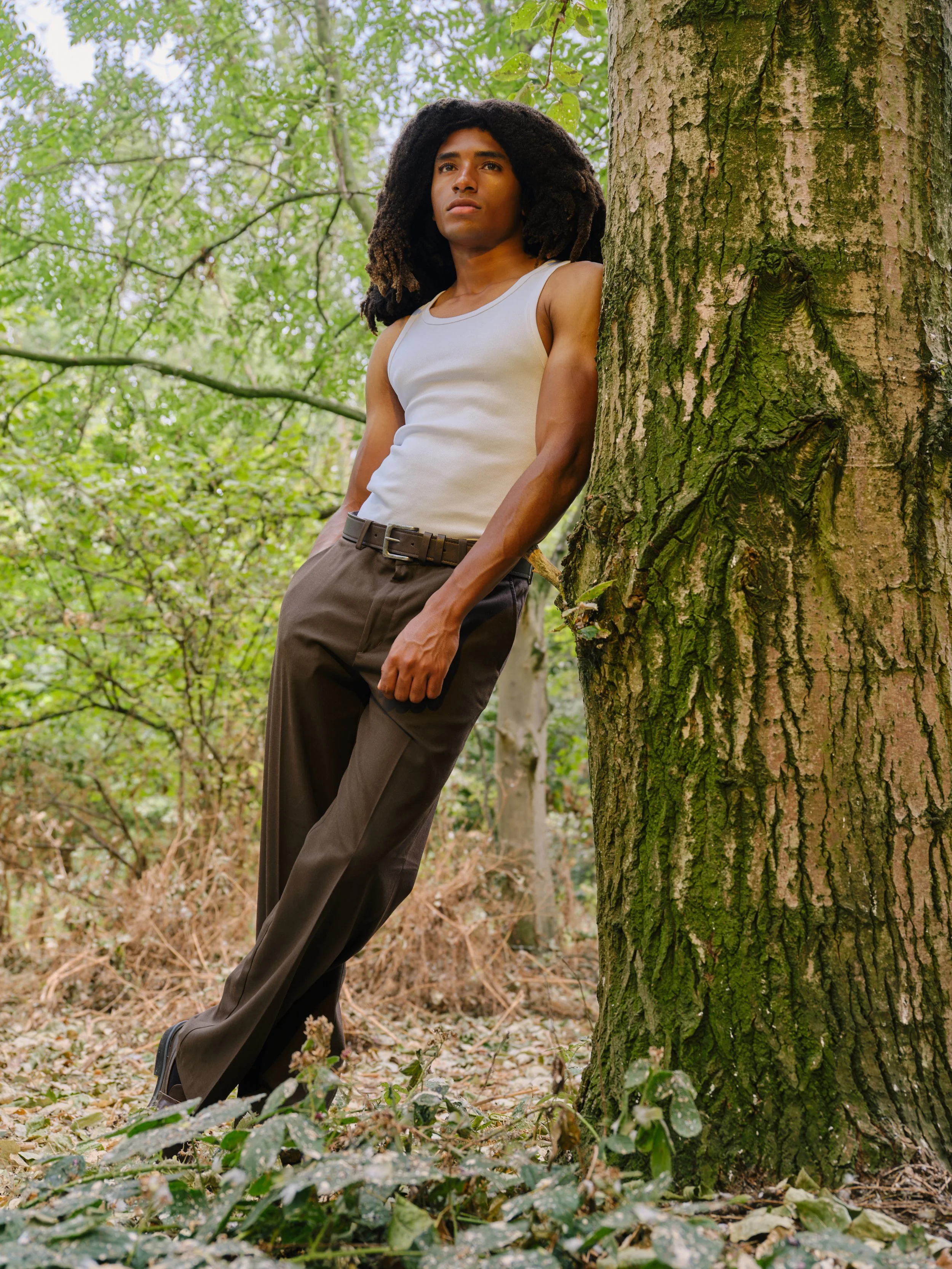 A young man leaning against a tree in a forest. He is wearing a white sleeveless shirt and brown pants, with long, curly black hair.